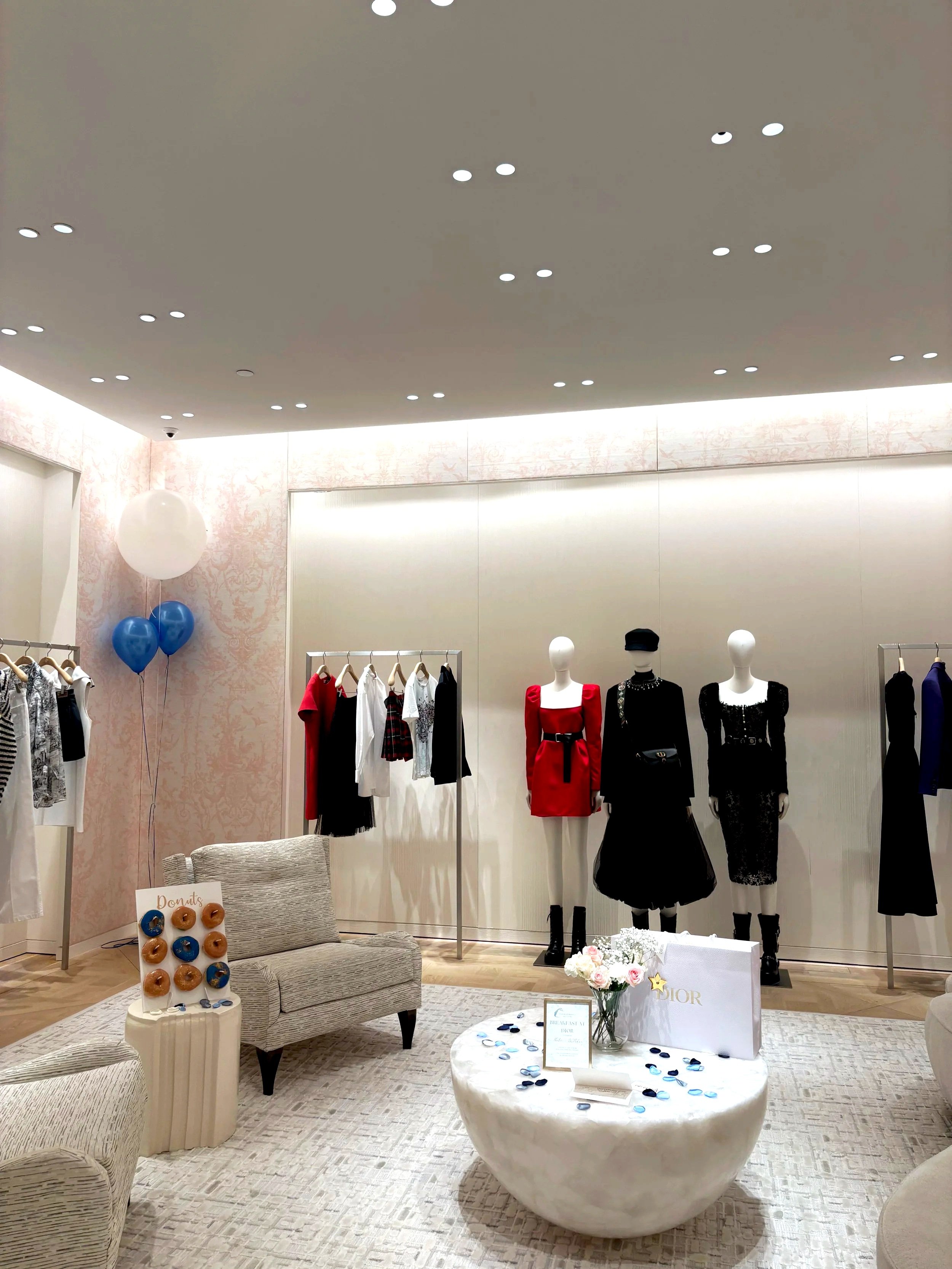 A boutique clothing store display with mannequins dressed in fashionable outfits, clothing racks with various garments, a small seating area with a sofa and armchair, a round coffee table with flowers and promotional items, and blue and white balloons in the corner.