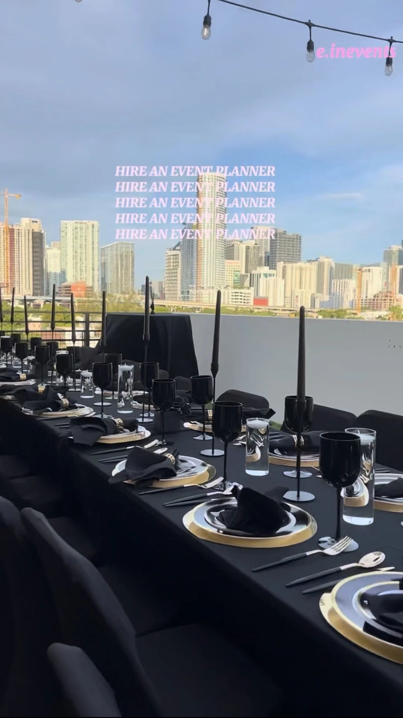 Set dining table with black tablecloth, black napkins, black and gold chargers, black glasses, and black candles, overlooking an urban city skyline through a window with string lights above.