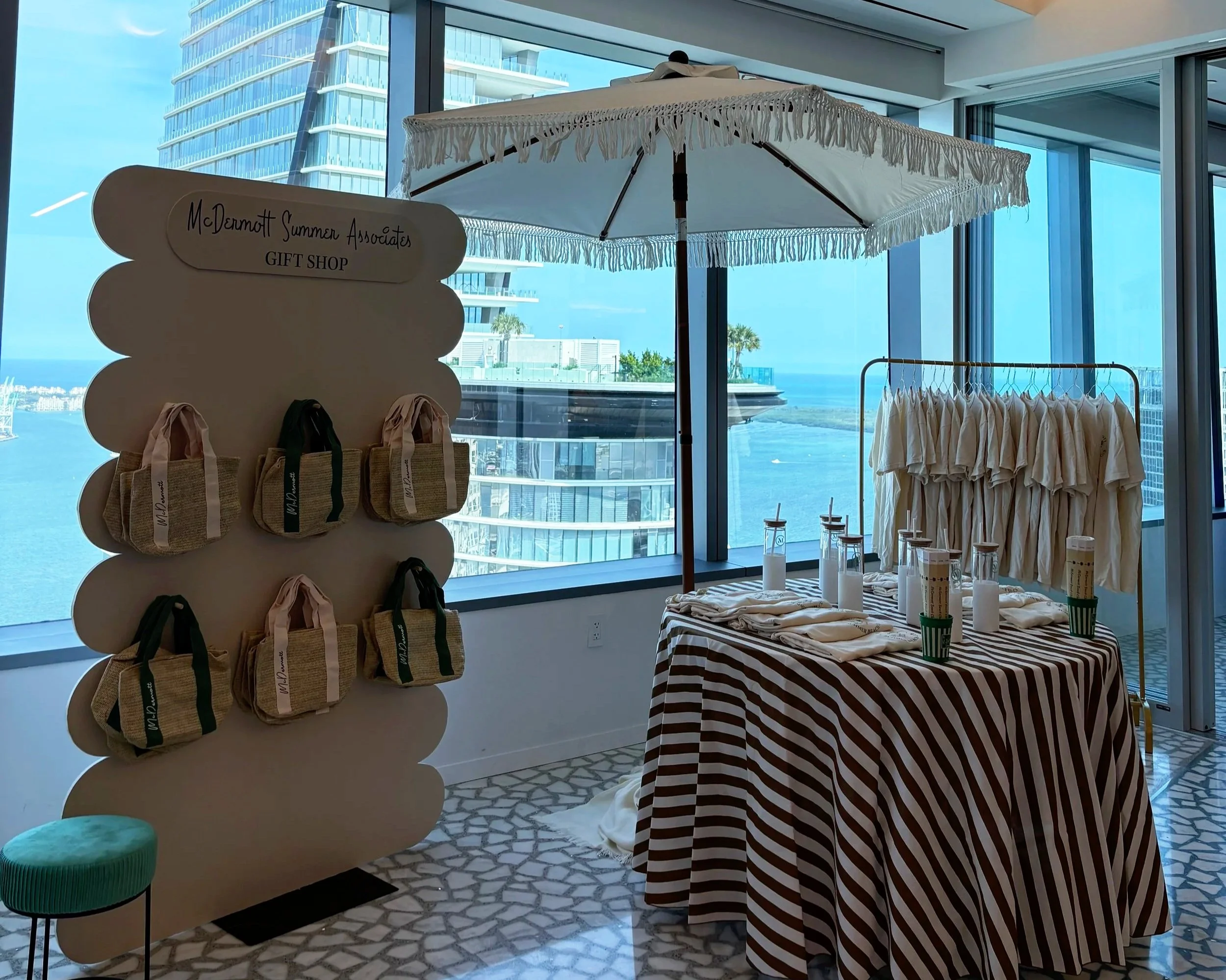 An indoor gift shop area with a view of a city skyline and water outside large windows. The shop features a display stand with small handbags, a table with candles, and hanging white t-shirts. There's a large white umbrella over the display table.