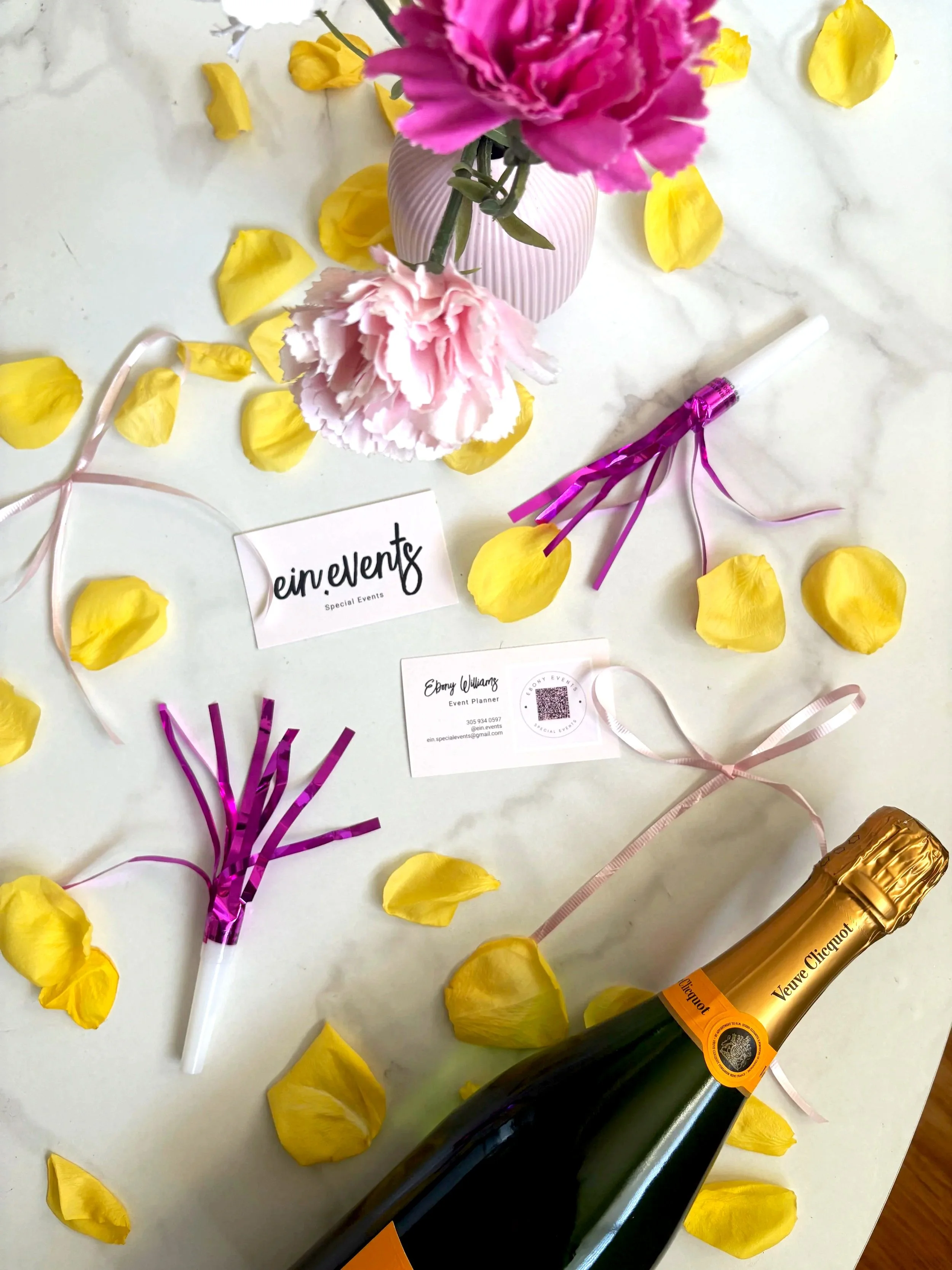 A celebration table decorated with pink and white flowers in a pink vase, yellow flower petals, a bottle of Veuve Clicquot champagne, pink and purple party blowers, and business cards for events.