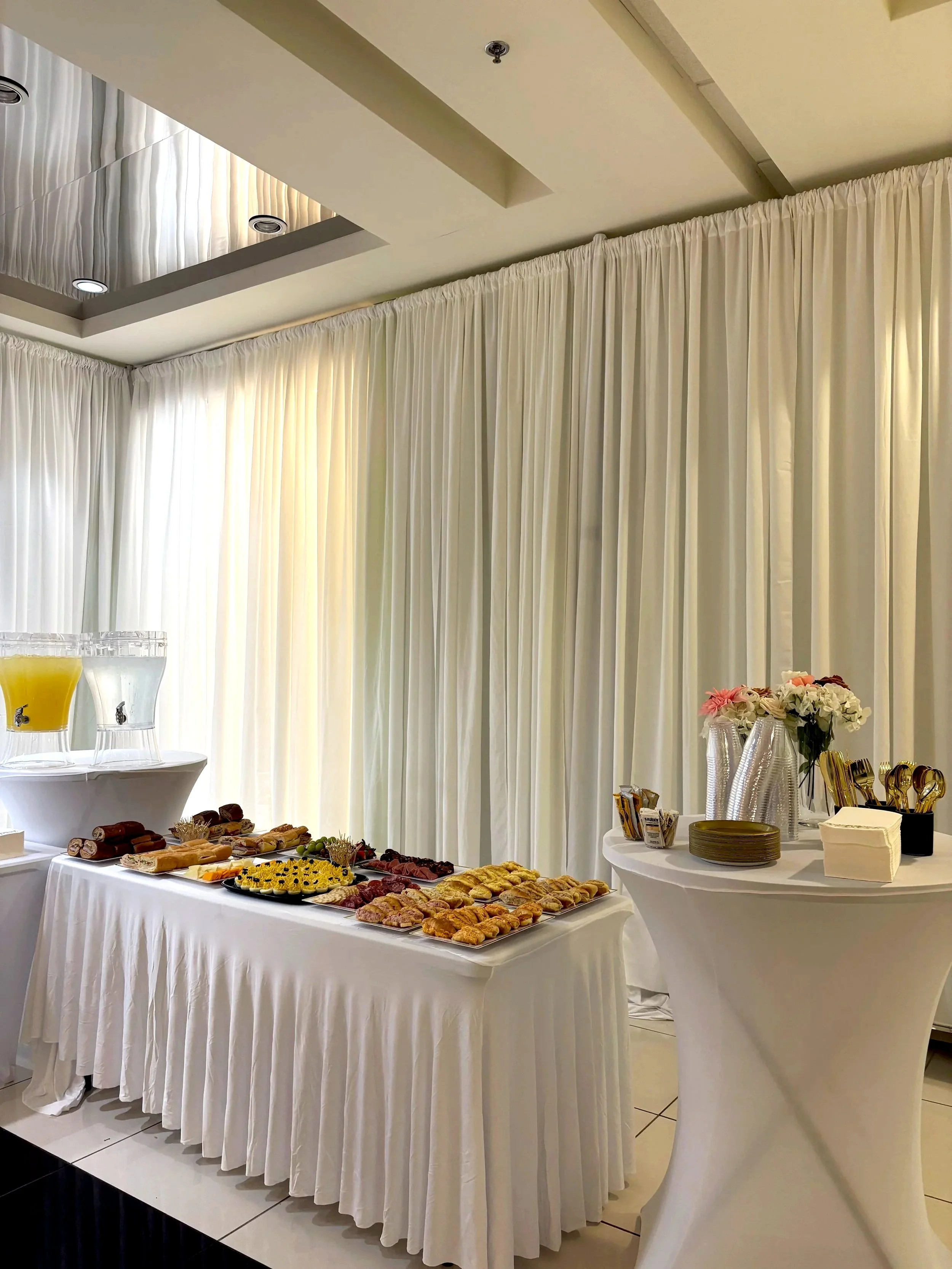Buffet table with pastries, fruit, and drinks in a room with white curtains and decorated tableware.