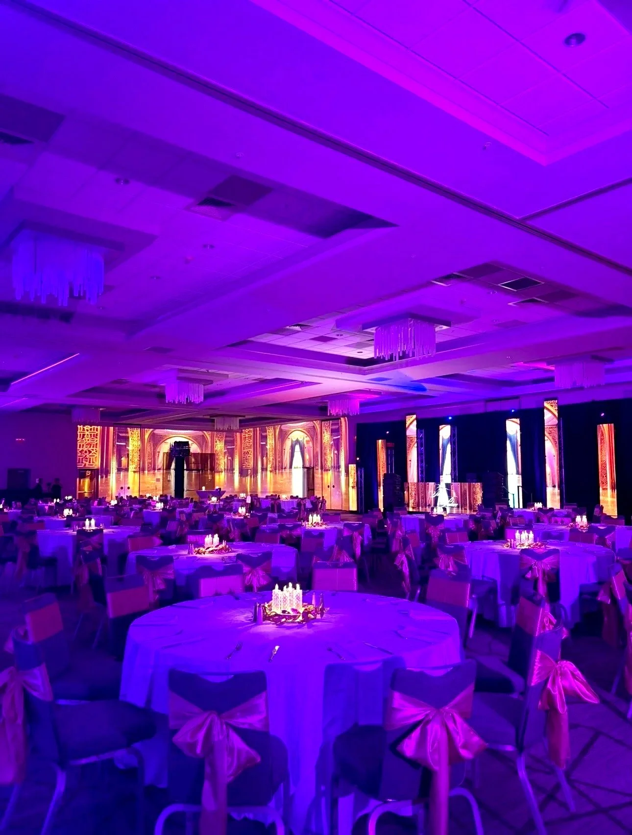 A banquet hall decorated for an event with purple lighting, elegant round tables with candles, chairs with pink bows, and a large illuminated backdrop on stage.