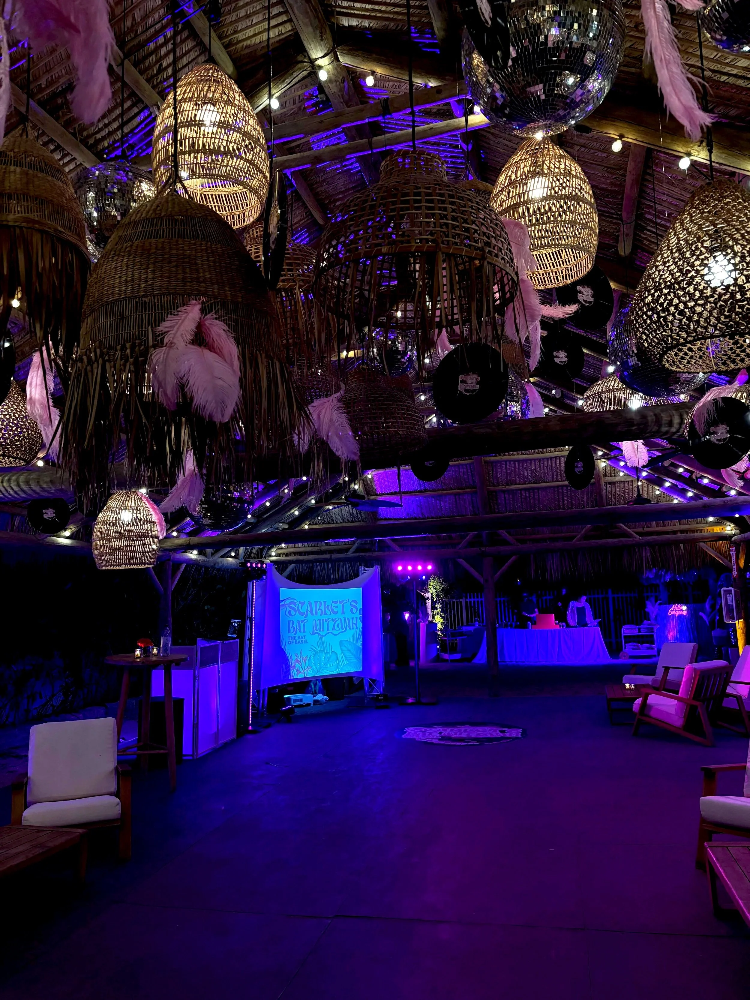 Decorative bamboo and wicker lamps with pink feathers hanging from the ceiling of a tropical-style event space, illuminated with purple lights.