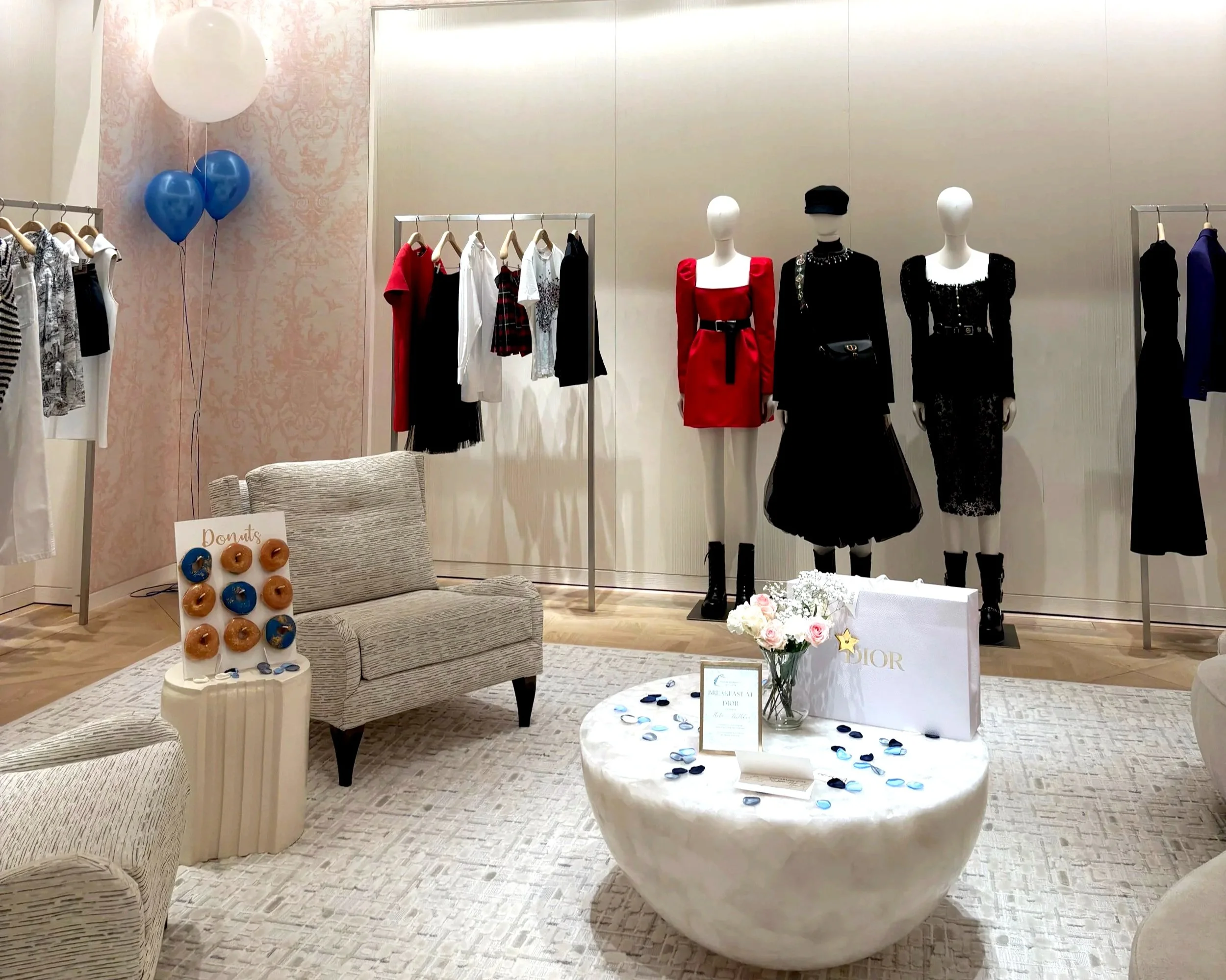 A boutique clothing store interior featuring mannequins dressed in fashionable outfits, clothing racks with various garments, a donut display with a 'Donuts' sign, and a table with Dior shopping bags and promotional materials, decorated with flowers and blue confetti.