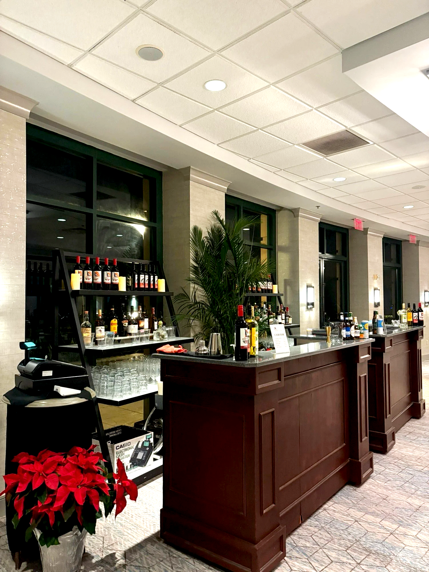 A bar setup in an indoor venue with a poinsettia plant in the foreground, dark wood counters, bottles of alcohol on racks and counters, glasses, and a cash register.