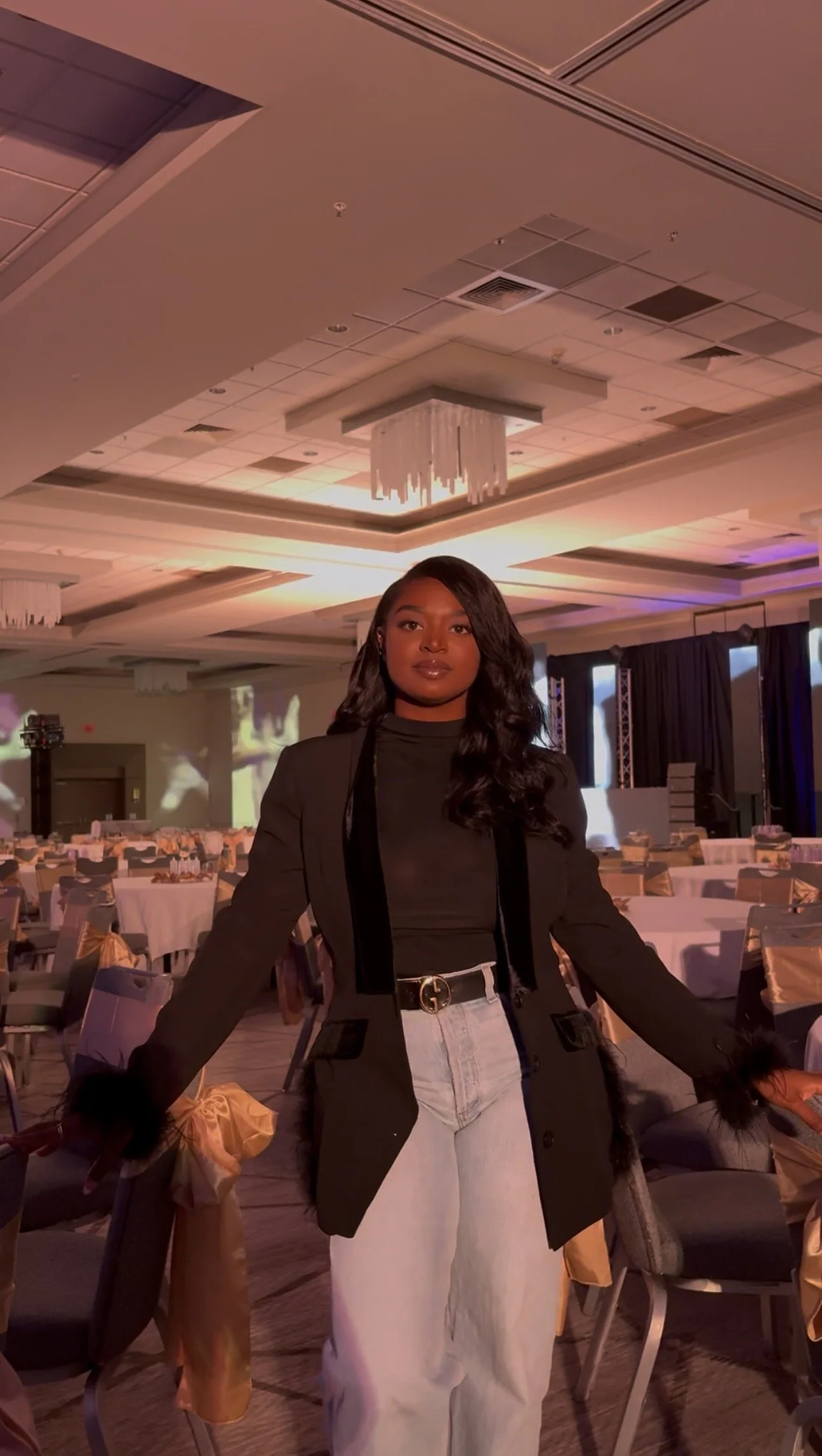 A woman standing in a large, decorated banquet hall with round tables and chairs, wearing a black blazer and light-colored jeans, with a chandelier and stage in the background.