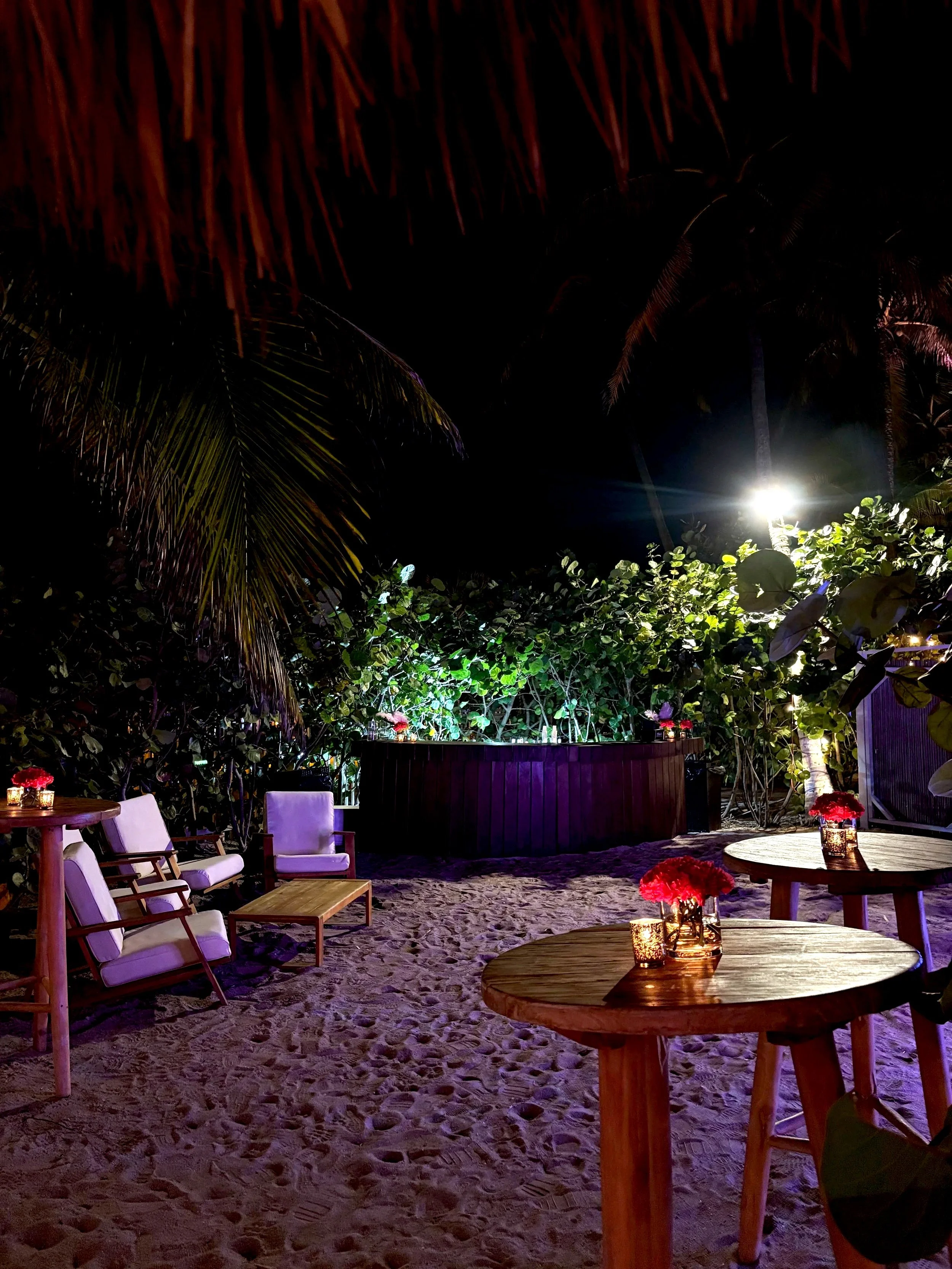 Nighttime outdoor setting with wooden tables, cushioned chairs, and lit candles, surrounded by lush greenery and palm trees, with a bright light in the background.