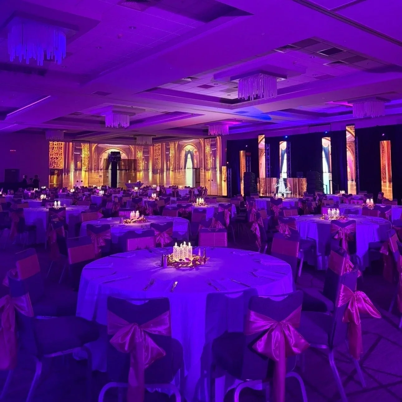 A bride with long red hair in a white dress is holding a sheer veil and dancing, while a groom with a bouquet of baby's breath flowers covering his face is standing next to her against a rustic wooden wall.