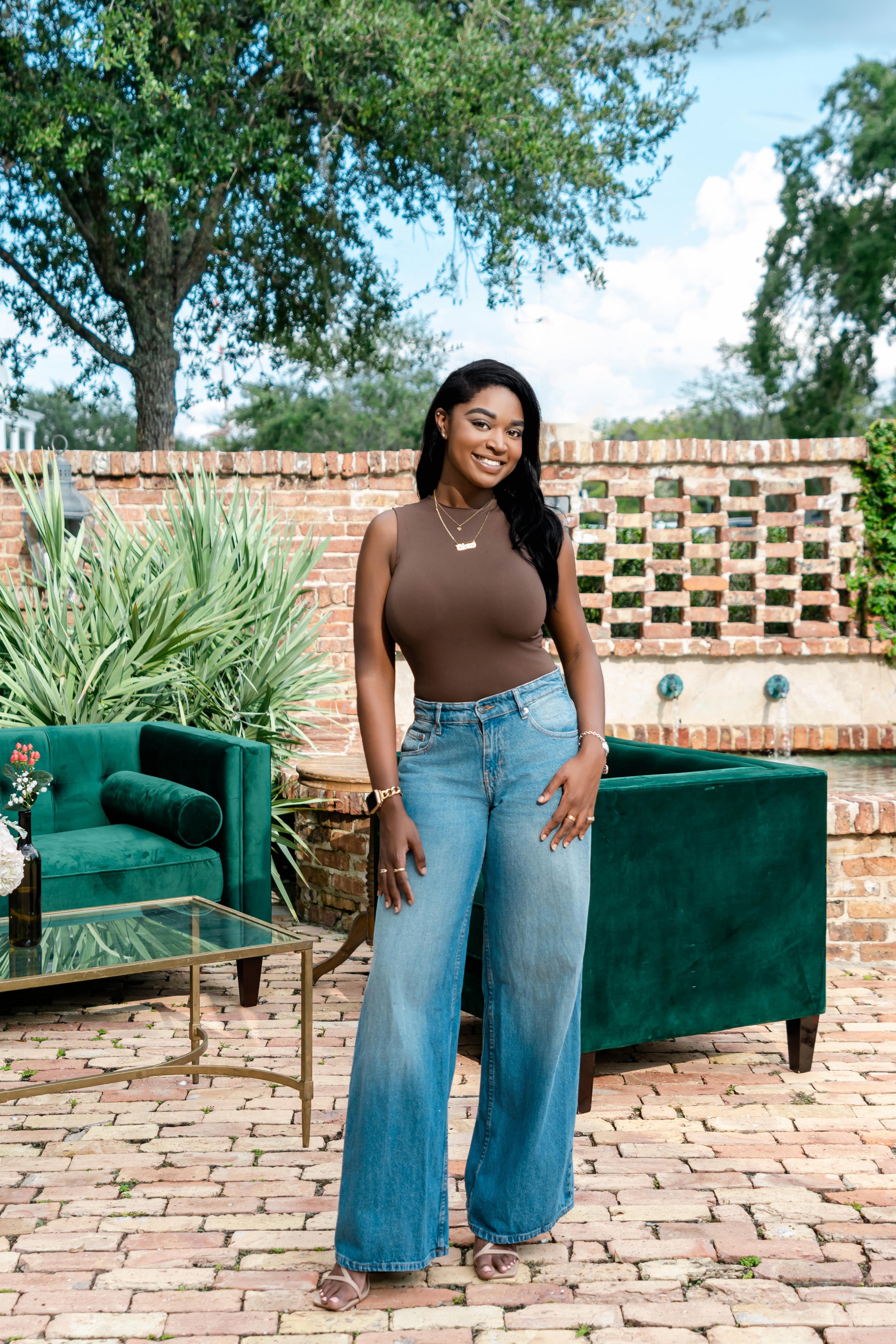 A woman standing outdoors on brick patio, smiling, wearing a sleeveless brown top and wide-leg blue jeans, with greenery and brick wall in the background.