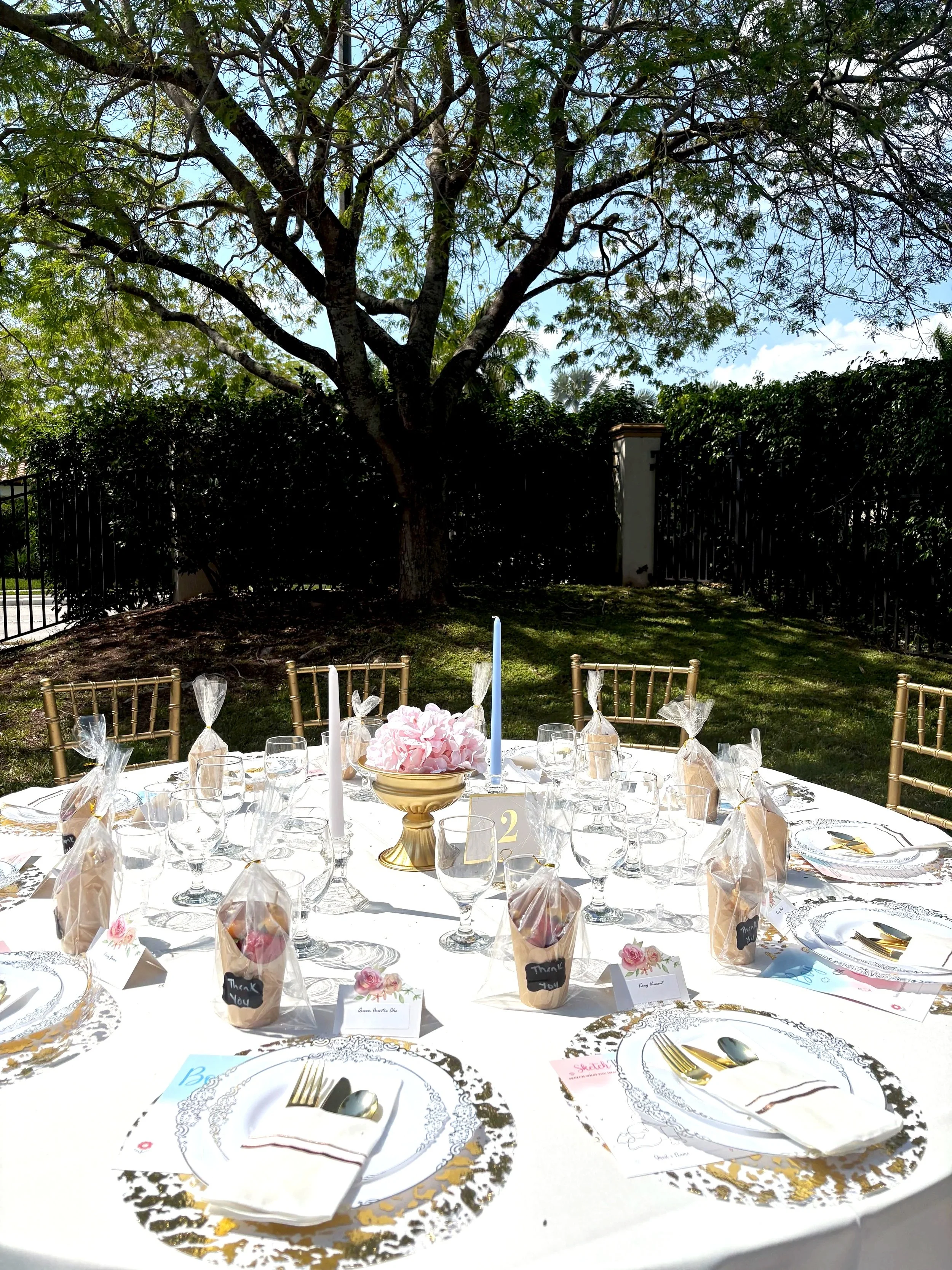 Decorated outdoor table set for a celebration, with elegant gold and white tableware, wrapped favor bags, a centerpiece of pink flowers, and a tall blue candle, under a large tree with green leaves.