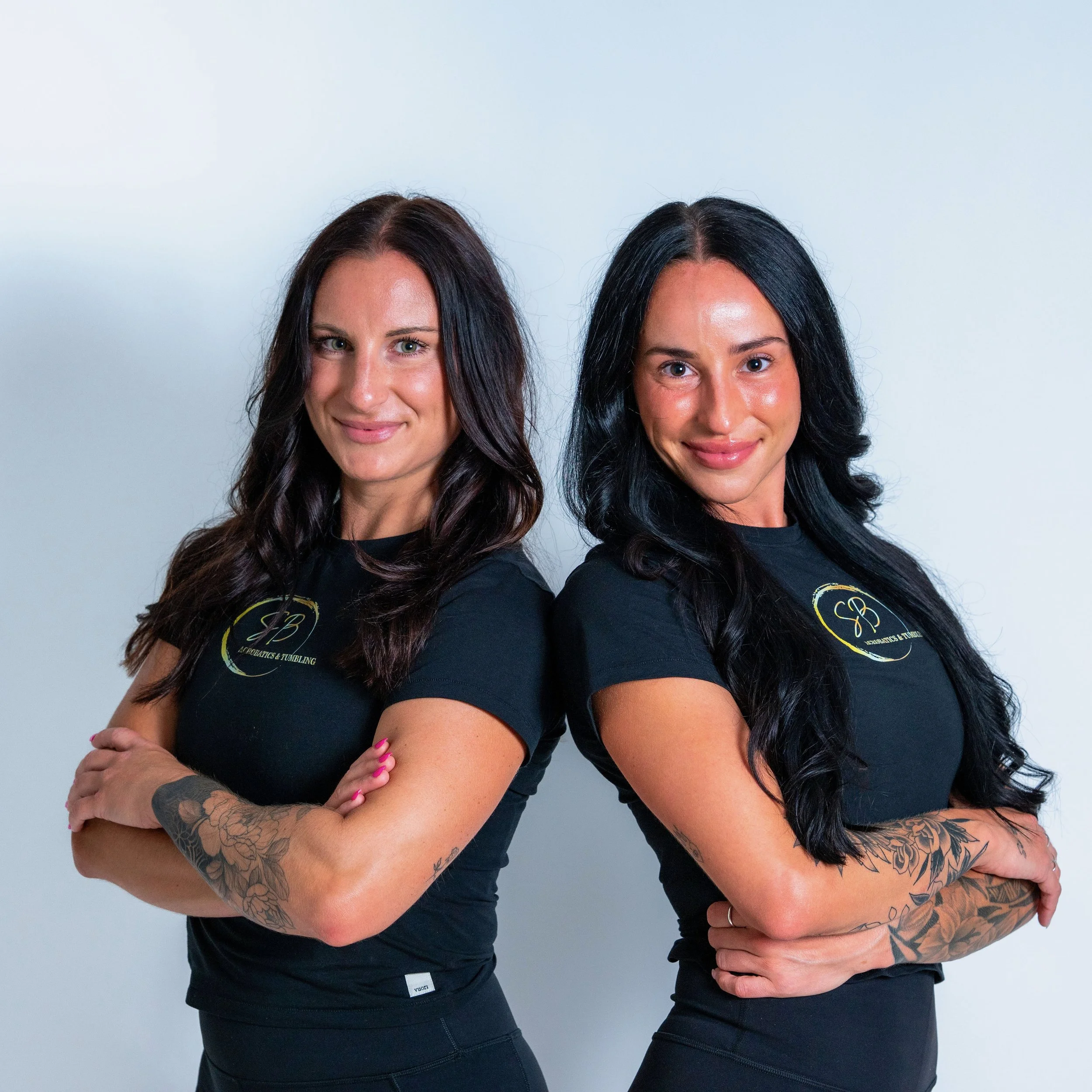 Two women standing with arms crossed, smiling, wearing black T-shirts with a logo, against a plain white background.