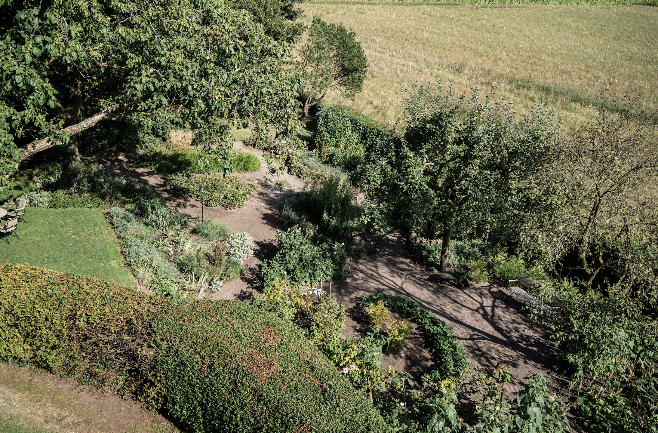 Vogelperspektive auf einen Garten mit Bäumen, Sträuchern, Wegen und Sitzgelegenheiten, umgeben von einer Wiese.