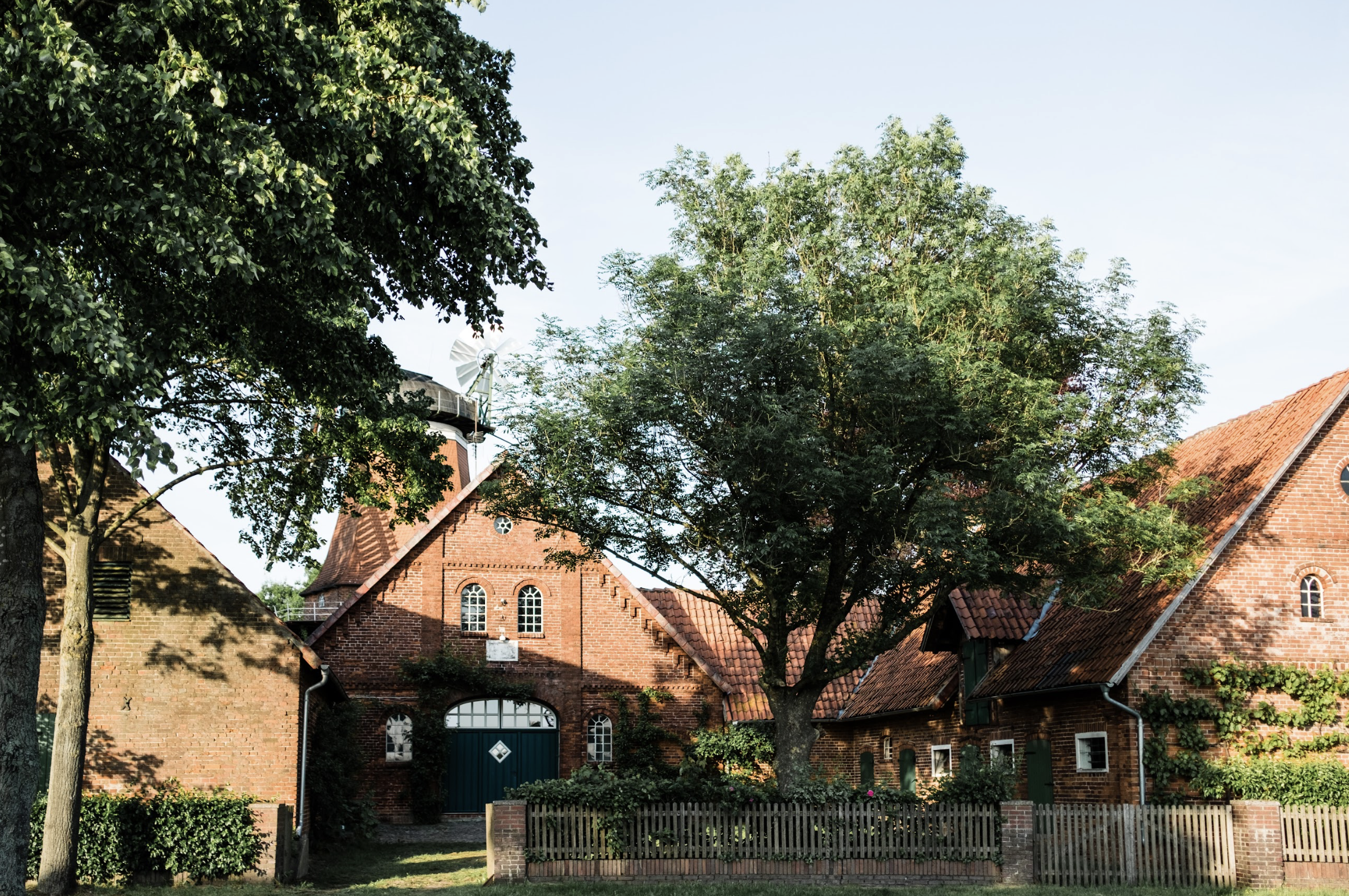 Ein landwirtschaftliches Gebäude mit roten Ziegeln, umgeben von Bäumen und einem Holz-Zaun, bei Sonnenschein.