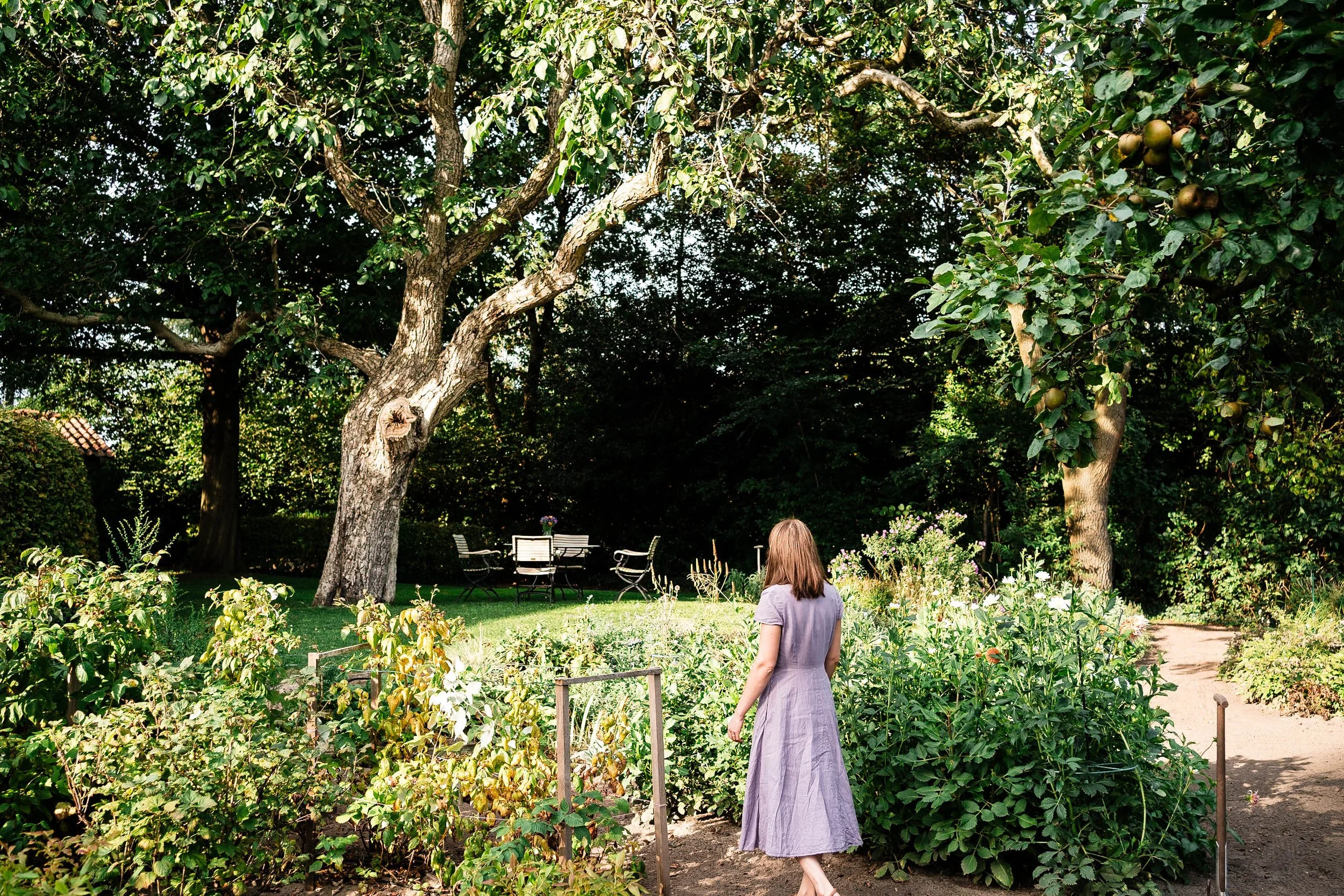 Eine Frau in einem lilafarbenen Kleid spaziert durch einen Garten mit Blumen und Bäumen, im Hintergrund sind Gartenmöbel.