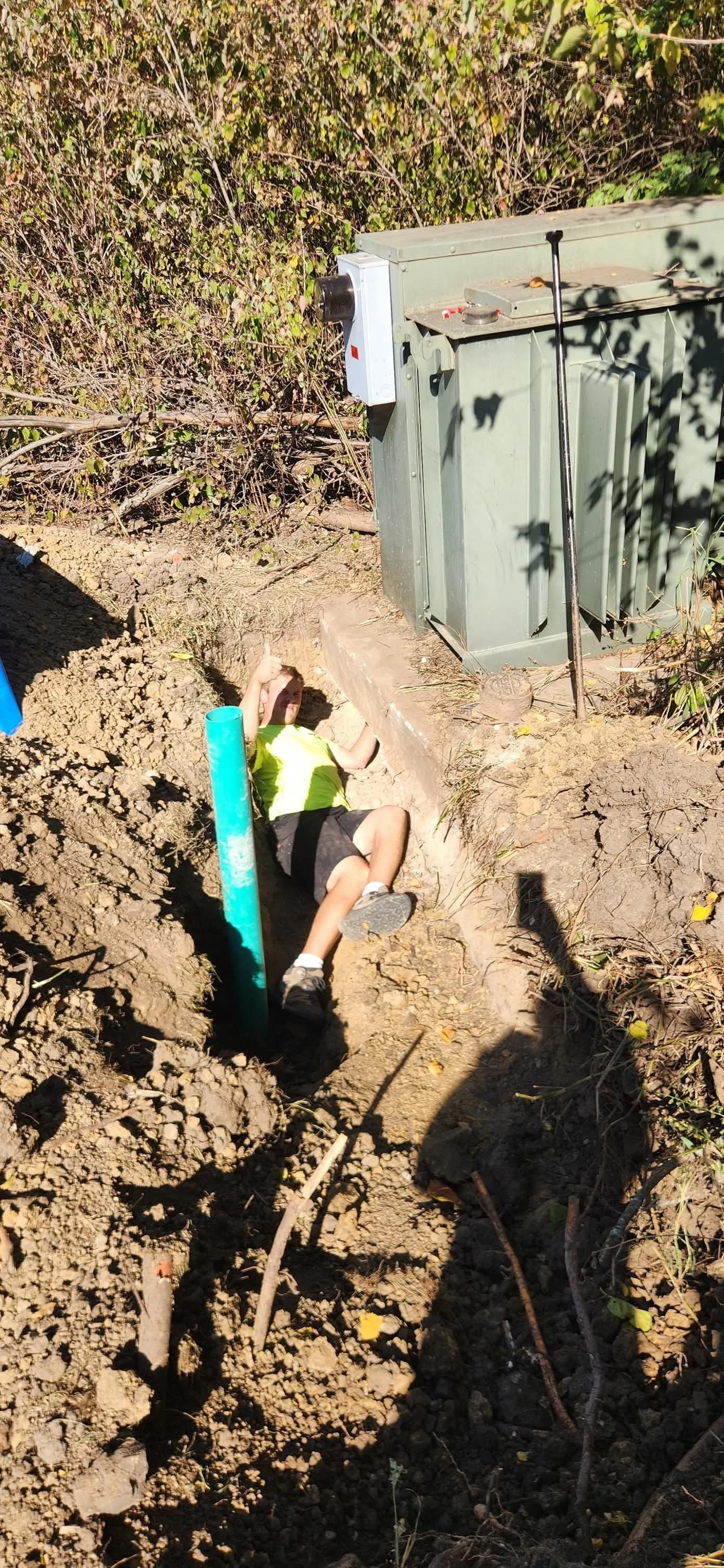 A person lying in a dirt trench with an open side, wearing a neon yellow shirt, black shorts, and white shoes, beside an electrical box and a green pole, with bushes and trees in the background.