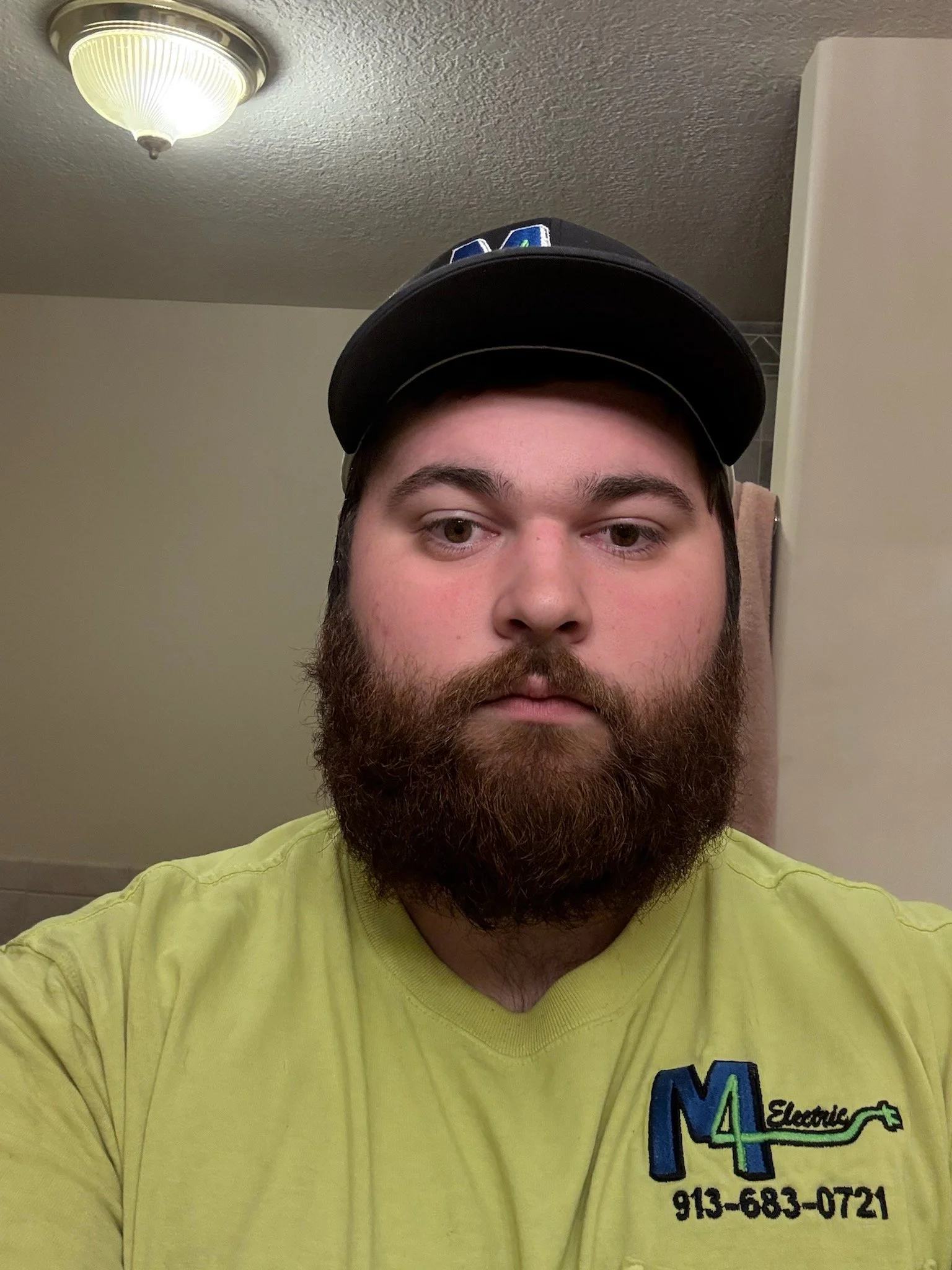 A man with a full beard and mustache wearing a black cap and a yellow shirt with a company logo and phone number on it, standing in a bathroom with a ceiling light fixture and a towel hanging in the background.