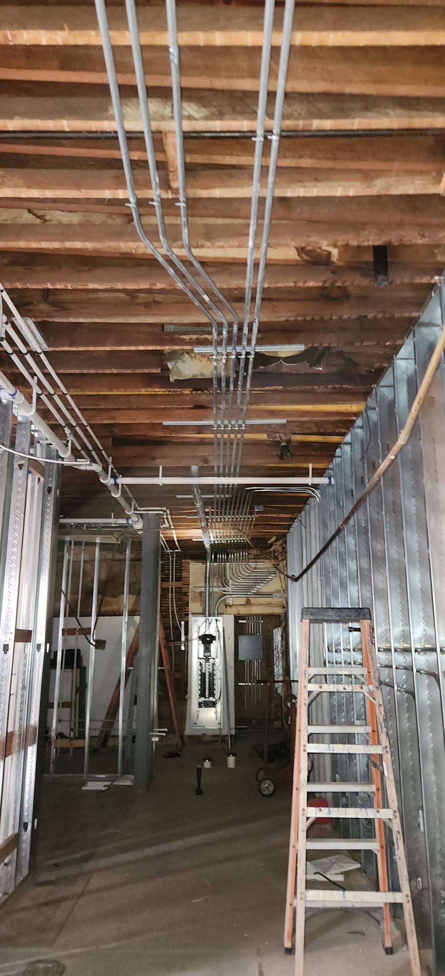 Electrical wiring and metal framing inside a building under construction with a wooden ceiling and with a ladder leaning against the wall.