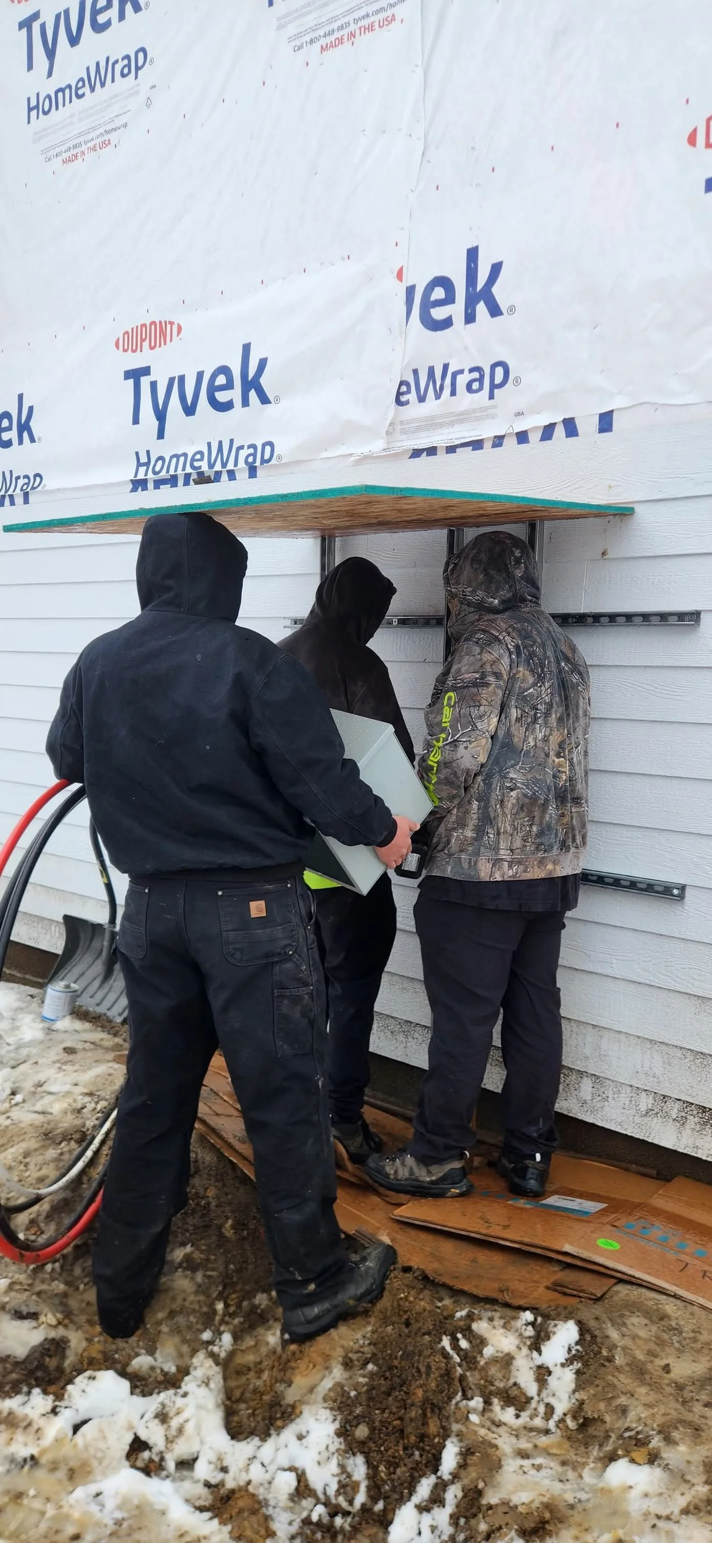 Three people working on the exterior of a house, installing or repairing something beneath the siding, with some snow and dirt on the ground.