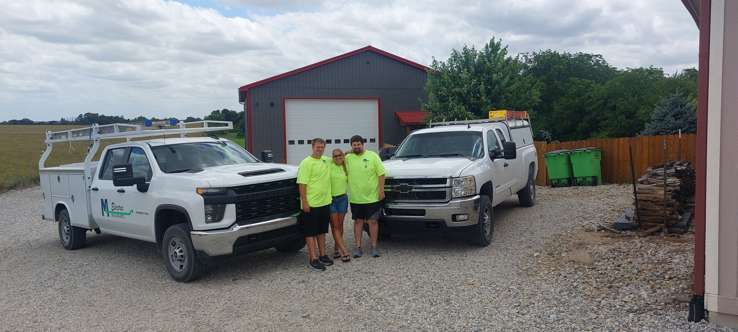 Three people stand together outdoors near two utility trucks, with a red and gray barn and trees in the background. The people are wearing bright neon green shirts.