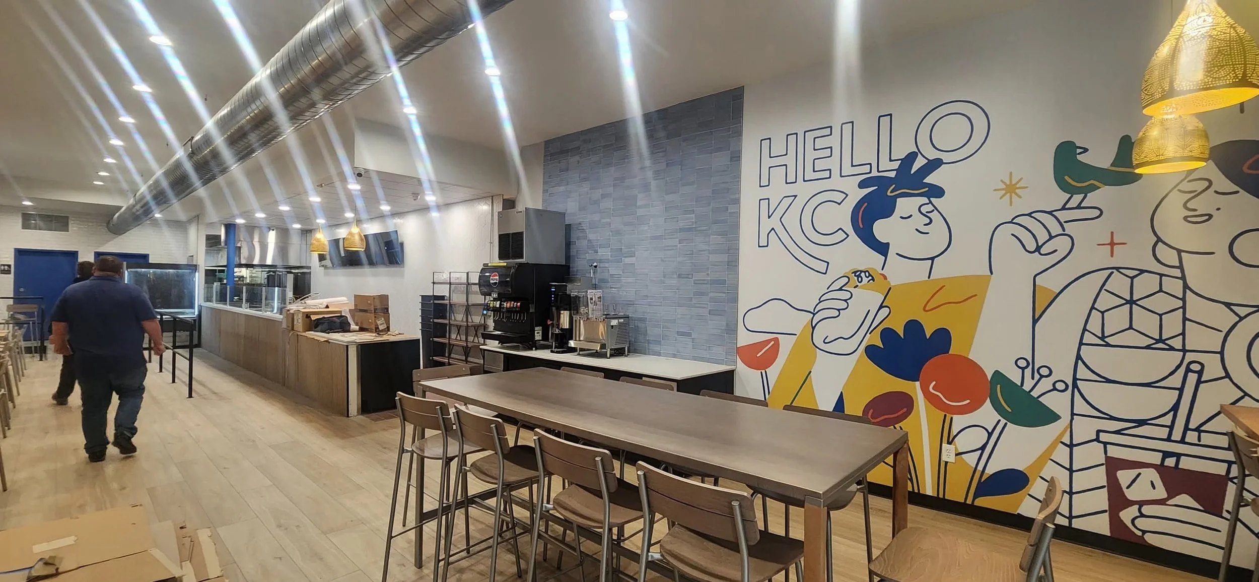 Interior of a restaurant or cafe with a large colorful mural on one wall featuring cartoon characters and the text 'HELLO KC'. There are tables and chairs, a beverage station, and a large duct running along the ceiling.