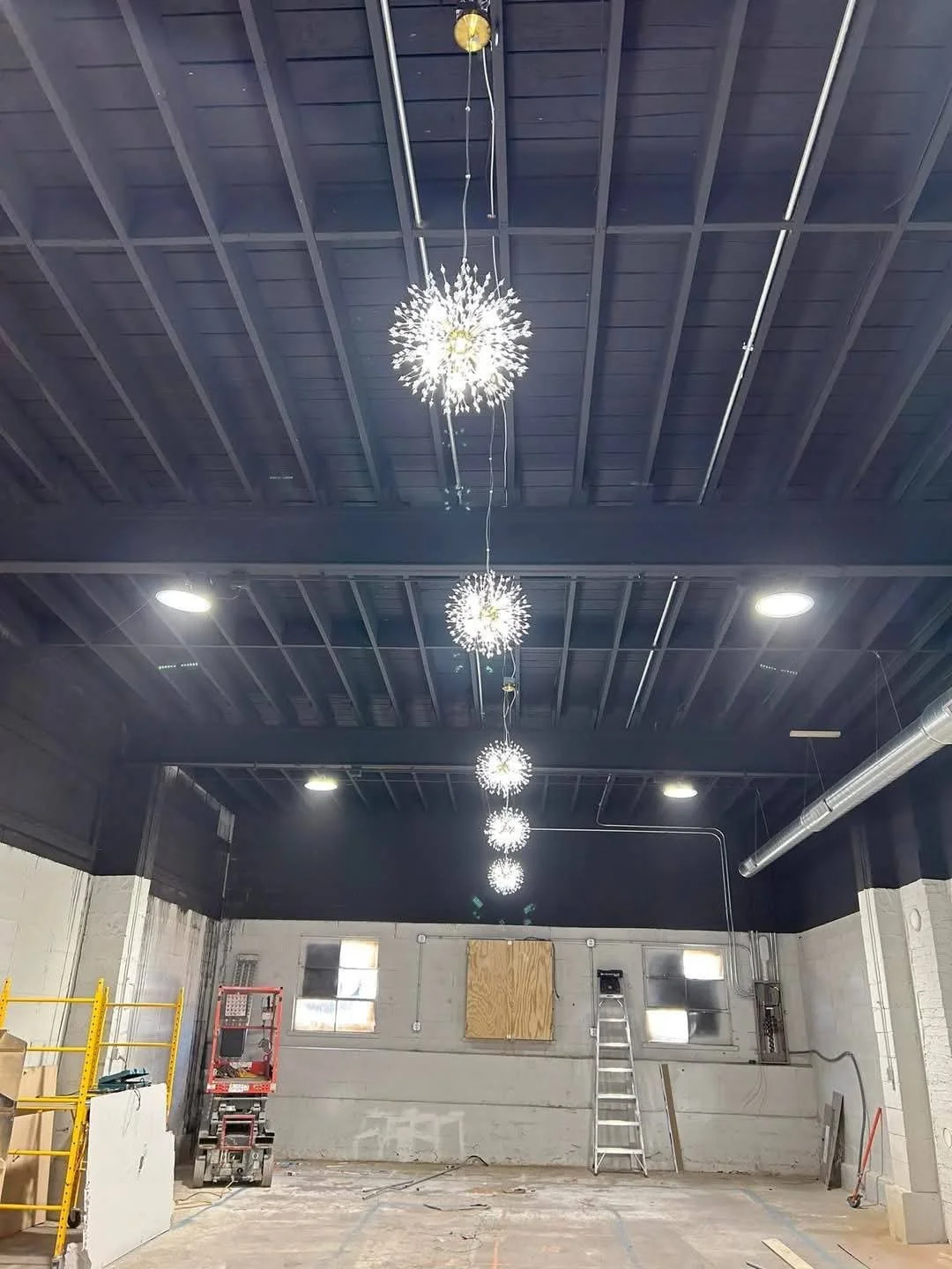 Interior of an industrial space under construction with black ceiling, hanging decorative light fixtures, a ladder, scaffolding, and construction tools.