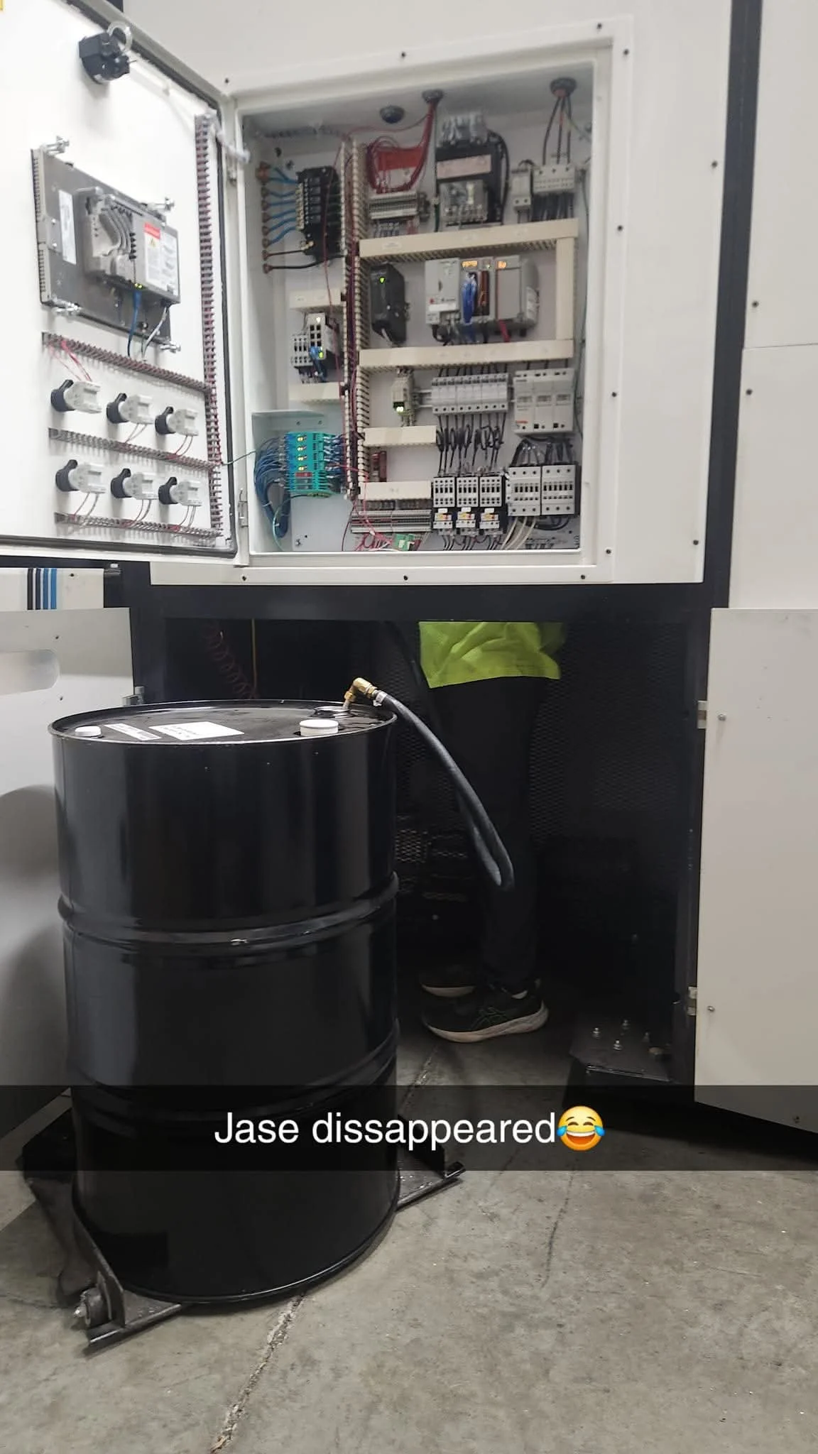 Open electrical control panel with wiring and components, and a large black drum on the floor connected with a hose. Person's legs and shoes visible behind the drum.