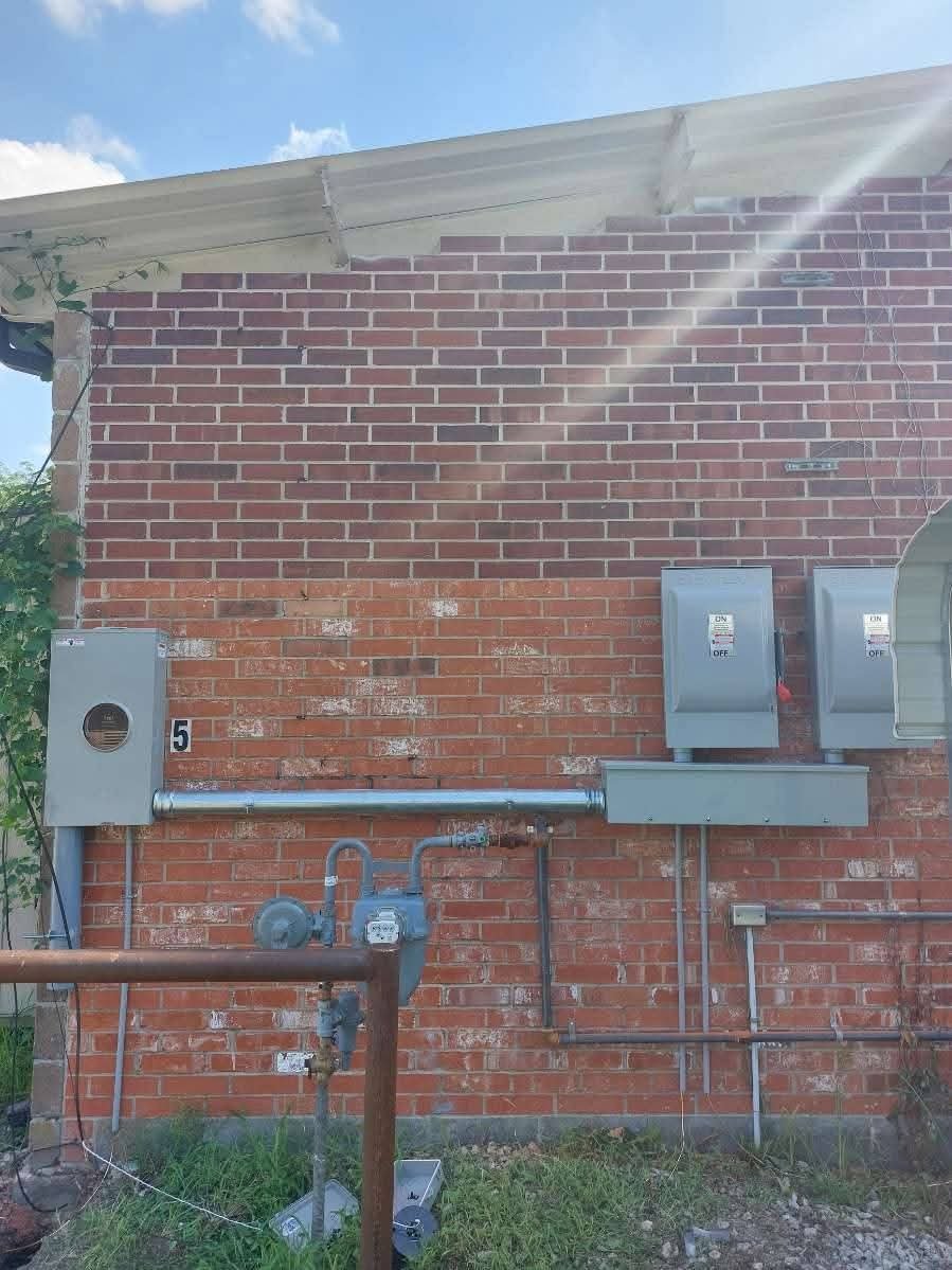 Exterior brick wall with electrical meters and boxes, some piping, and a grassy area in front. Overcast sky visible at the top.
