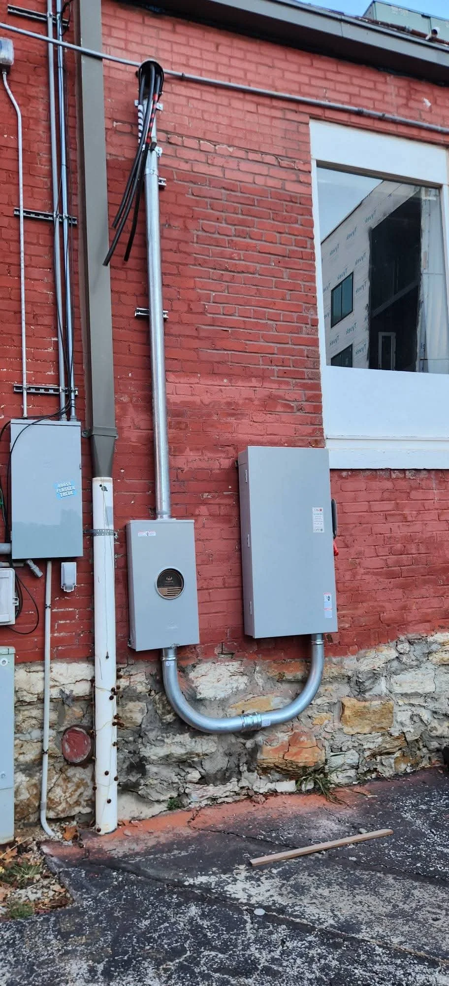 Exterior view of a brick building wall with electrical and utility boxes, conduits, and pipes attached.