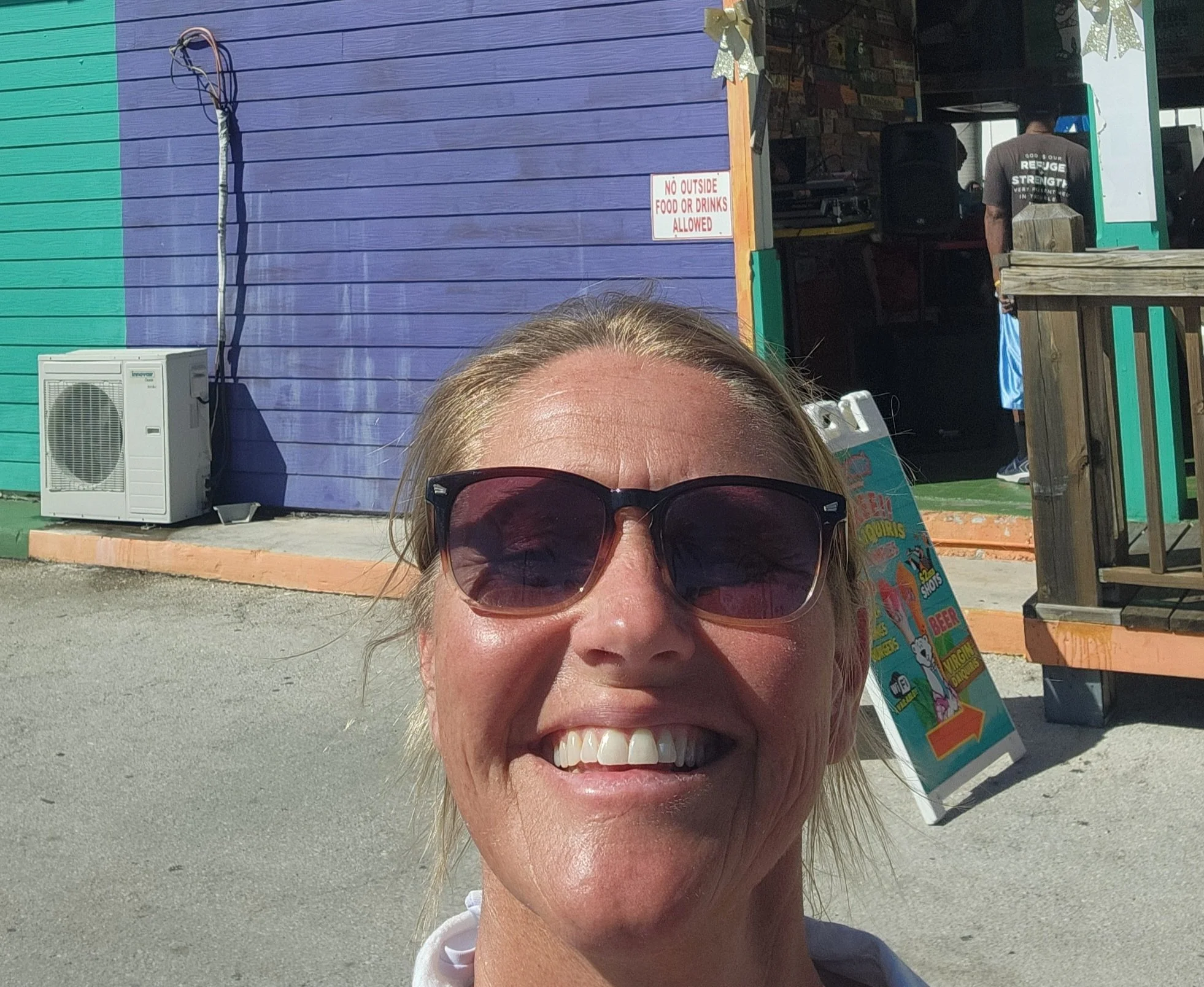 A woman wearing sunglasses and smiling in front of a colorful building with a sign that says 'No outside food or drinks allowed.'