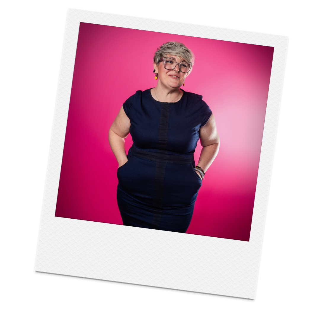A woman with short blonde hair, glasses, and earrings posing with hands in her pockets against a pink background.
