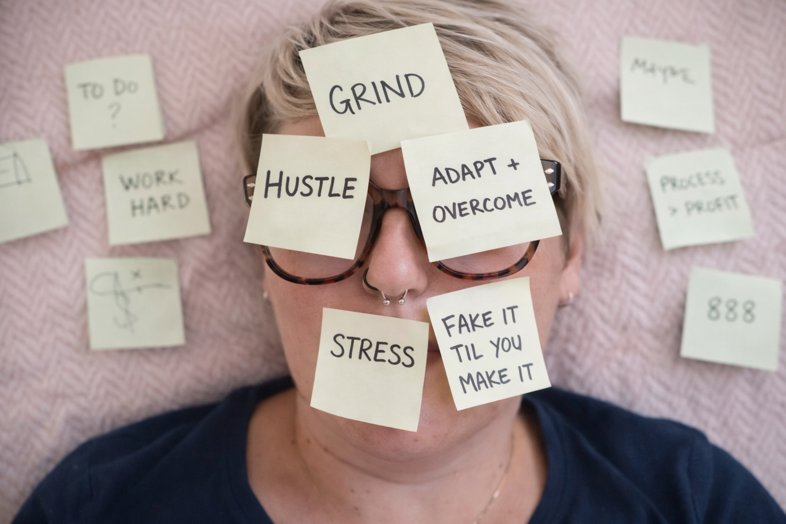 Ria lying on a soft surface with sticky notes covering her face, showing words like “hustle,” “grind,” “adapt and overcome,” “stress,” and “fake it till you make it,” representing pressure, overwork, and coping at work.