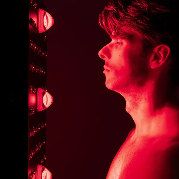A young man with tousled hair gazing at a wall of red medical or infrared lamps in a dimly lit room.