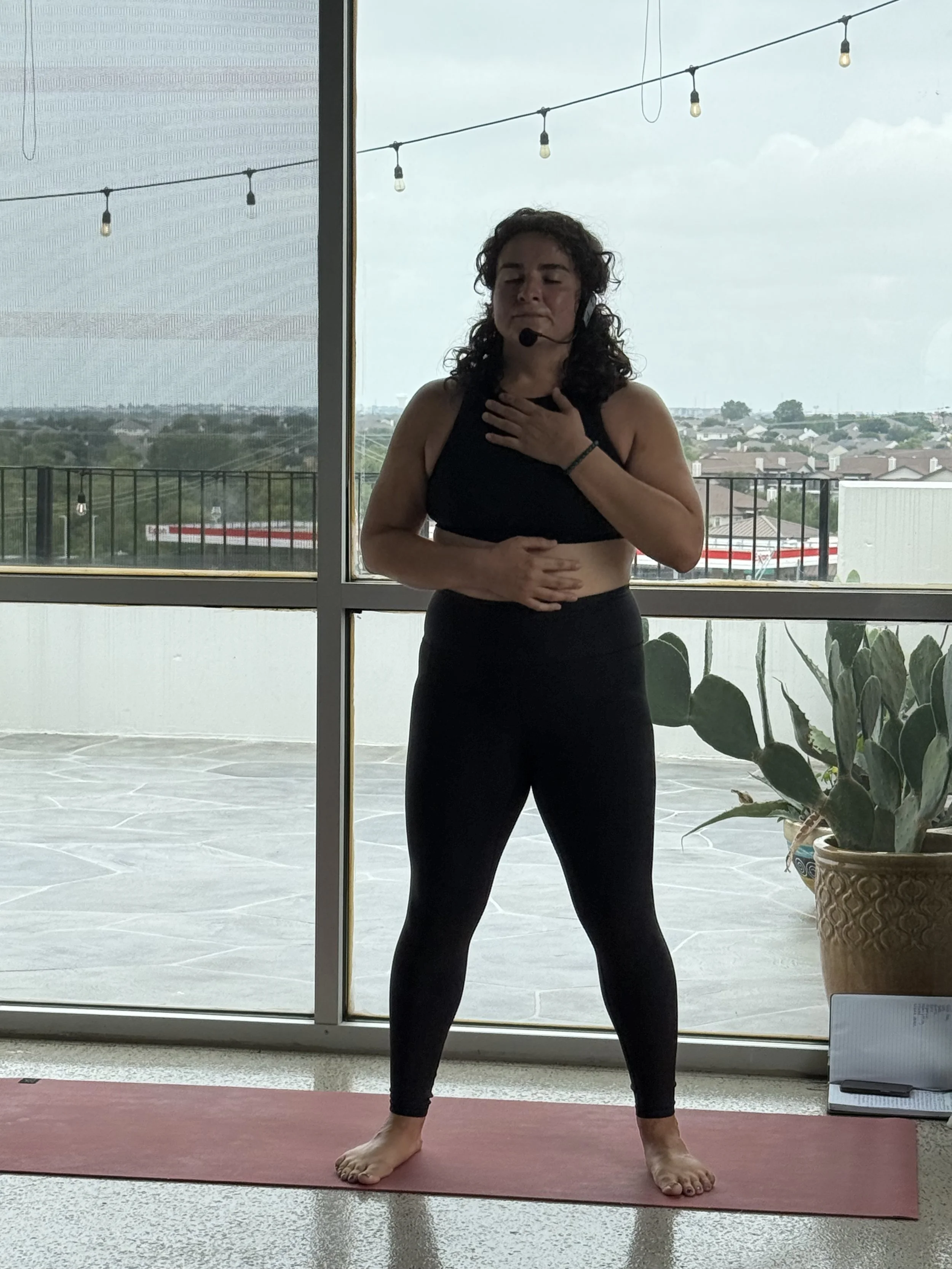 Woman with curly hair wearing a headset, black sports bra, and leggings, standing on a yoga mat with hand on her chest and stomach, in front of large windows showing an outdoor balcony and cloudy sky.