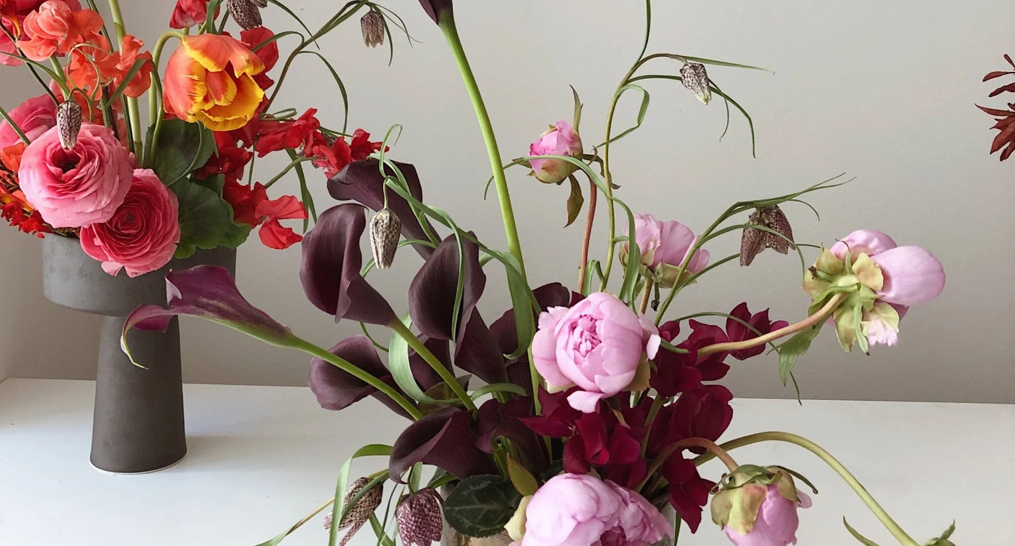 Two floral arrangements with roses, calla lilies, and other flowers in vases on a white surface against a neutral background.