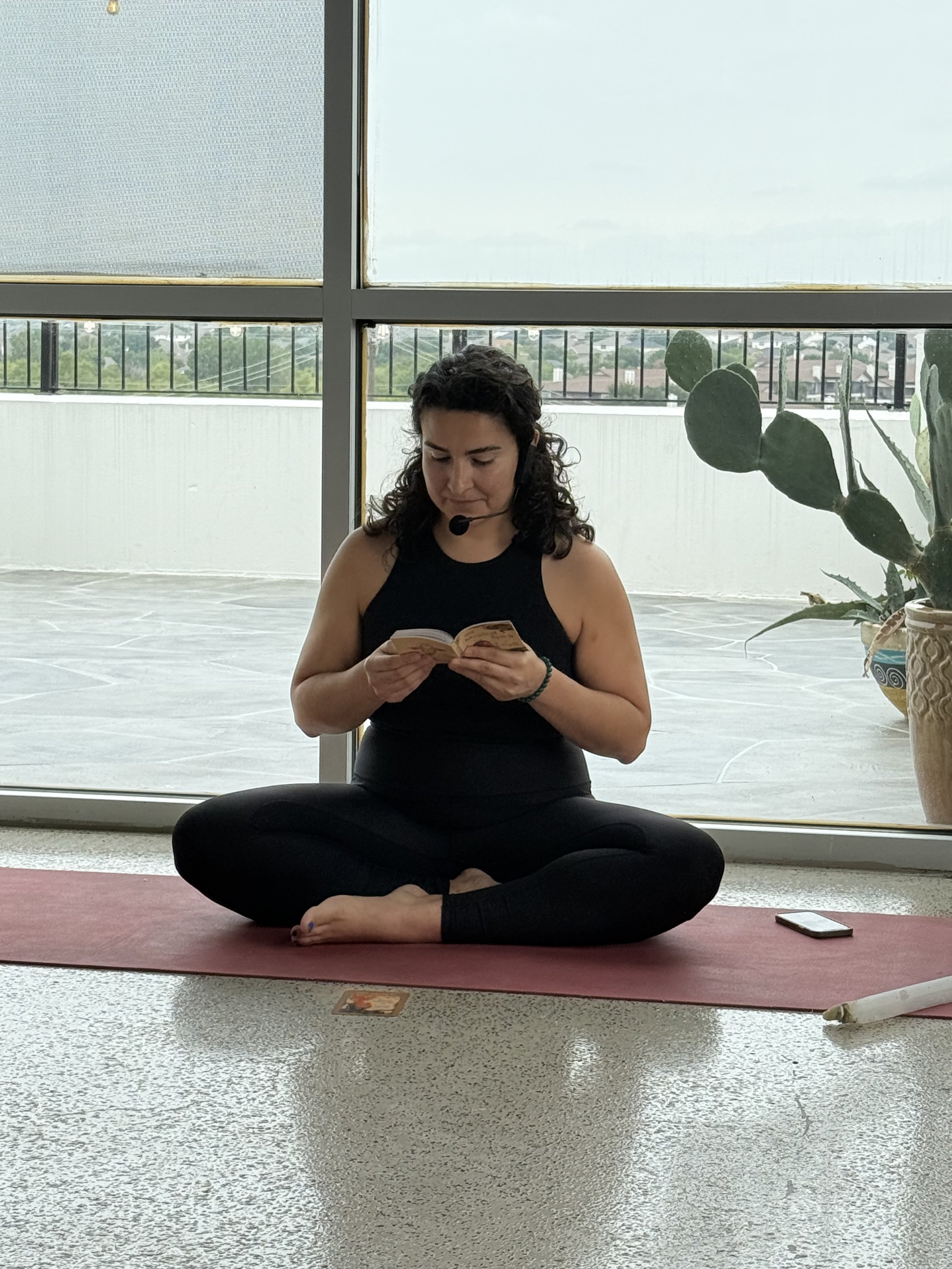 A woman sitting cross-legged on a yoga mat, holding a small book, wearing a headset microphone, in a room with large windows and potted plants.