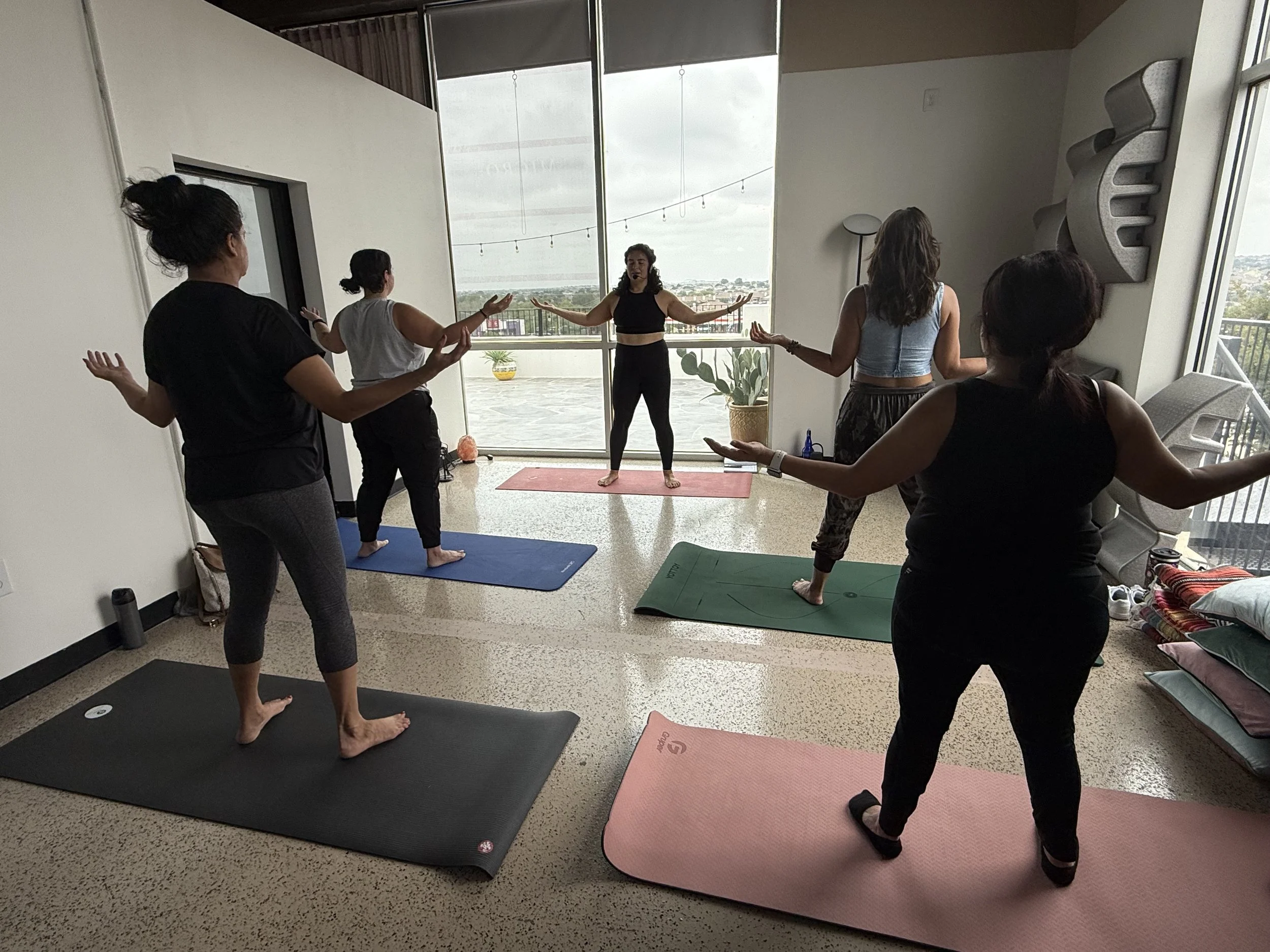 A group of five women practicing yoga on mats in a bright room with large windows and an outdoor balcony. The women are standing with arms raised or extended, following the instructor who is at the front, leading the session.