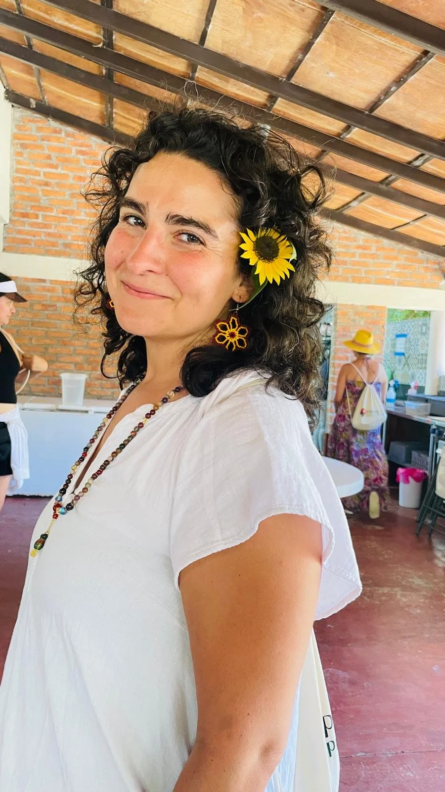 A woman with curly dark hair smiling, wearing sunflower earrings and a sunflower tucked behind her ear, dressed in a white top, standing indoors with a brick wall and ceiling beams visible in the background. There are other people in the background, including a woman in a hat and a colorful dress.