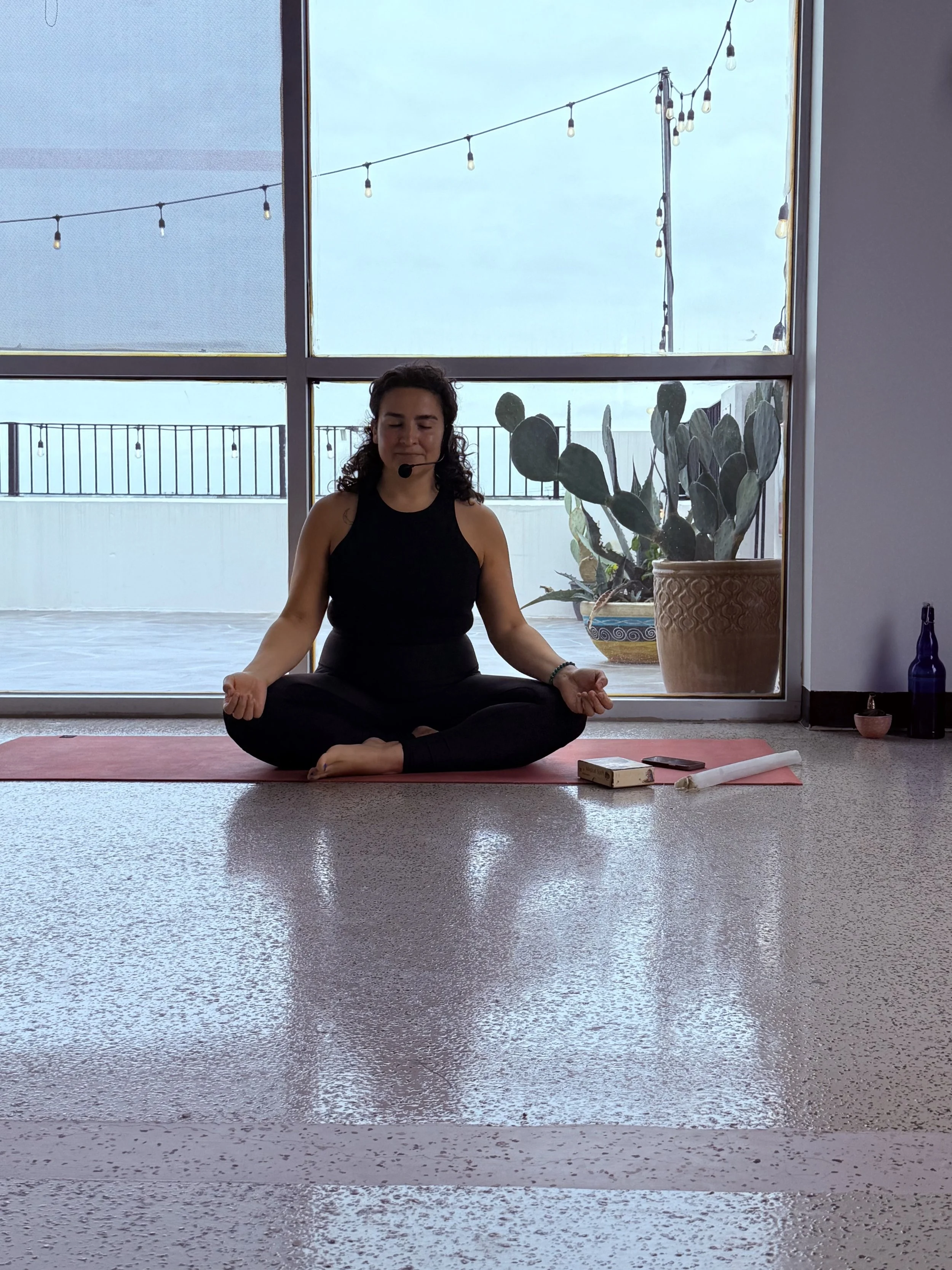 Woman practicing yoga in a seated meditation pose on a pink yoga mat in a room with large windows, potted cactus, and string lights outside.