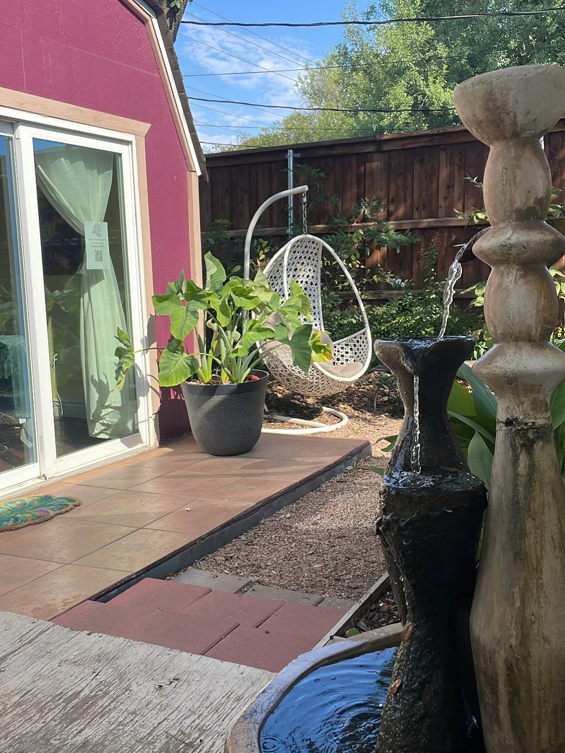 A backyard patio with a water fountain, potted plants, a hanging egg chair, and a sliding glass door leading into a house with a pink exterior