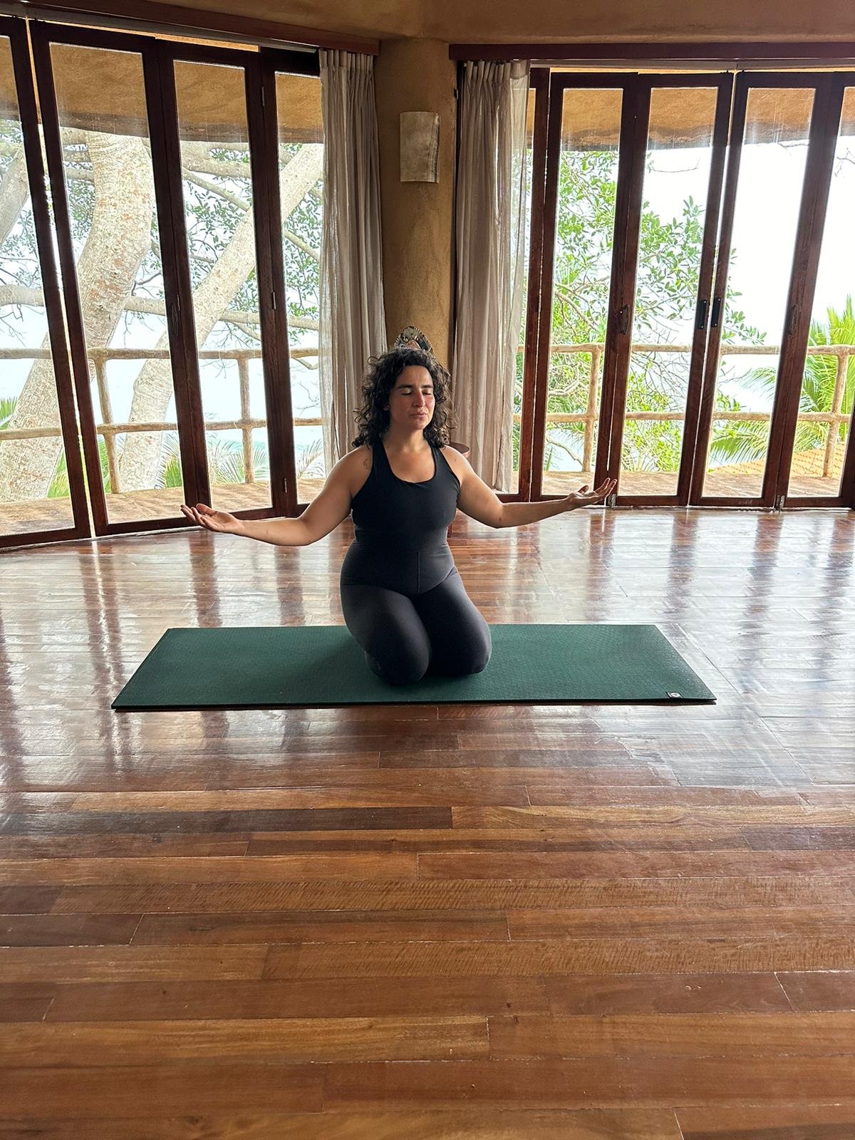 A woman practicing yoga indoors on a green mat, kneeling with arms extended and eyes closed, in front of large windows with curtains, overlooking trees and a balcony.