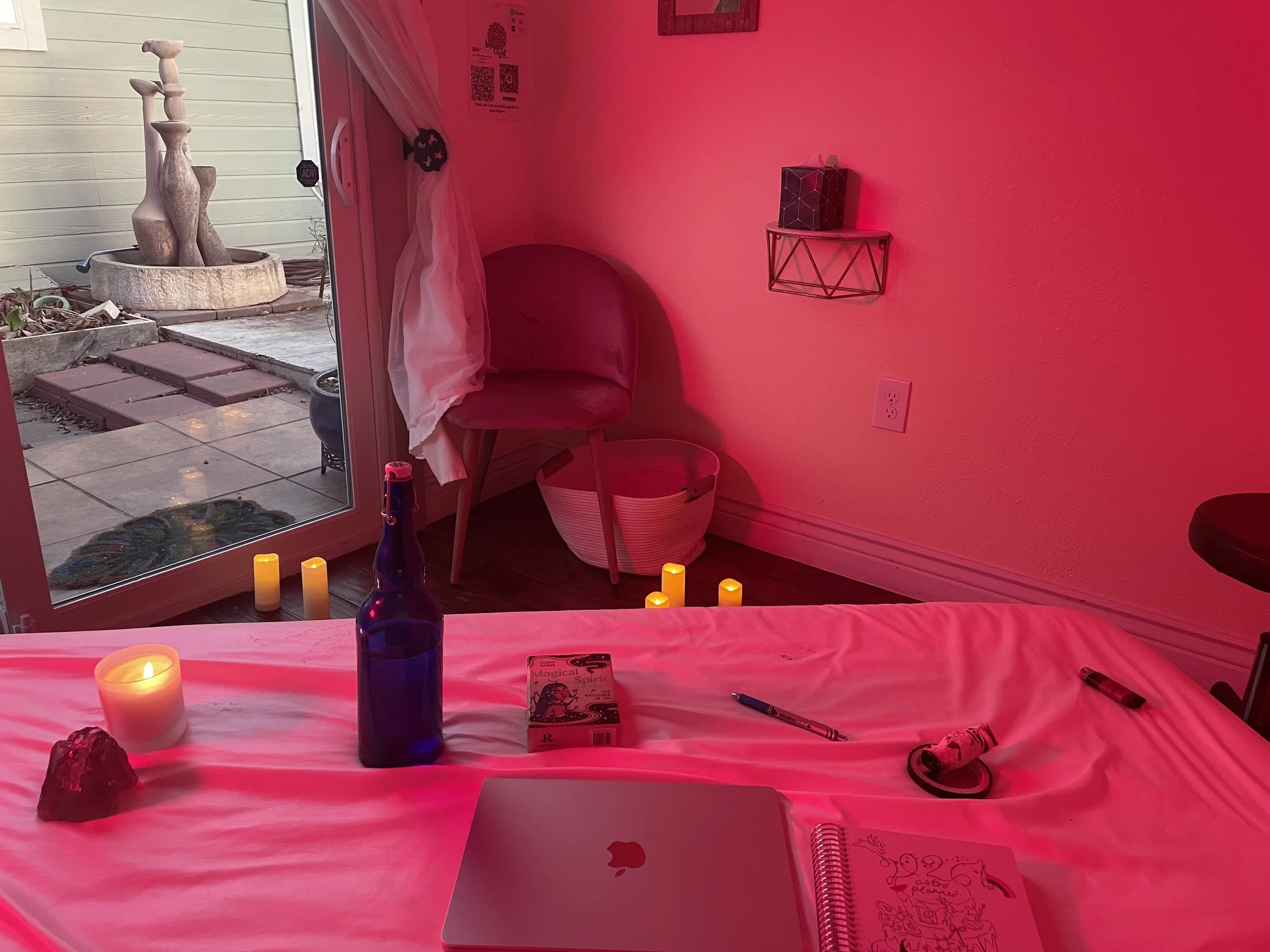 Indoor room with pink lighting, a table with objects including a laptop, a candle, a bottle, and paper, a chair with a white cloth, and candles on the floor. Outside through glass door is an outdoor patio with a fountain.