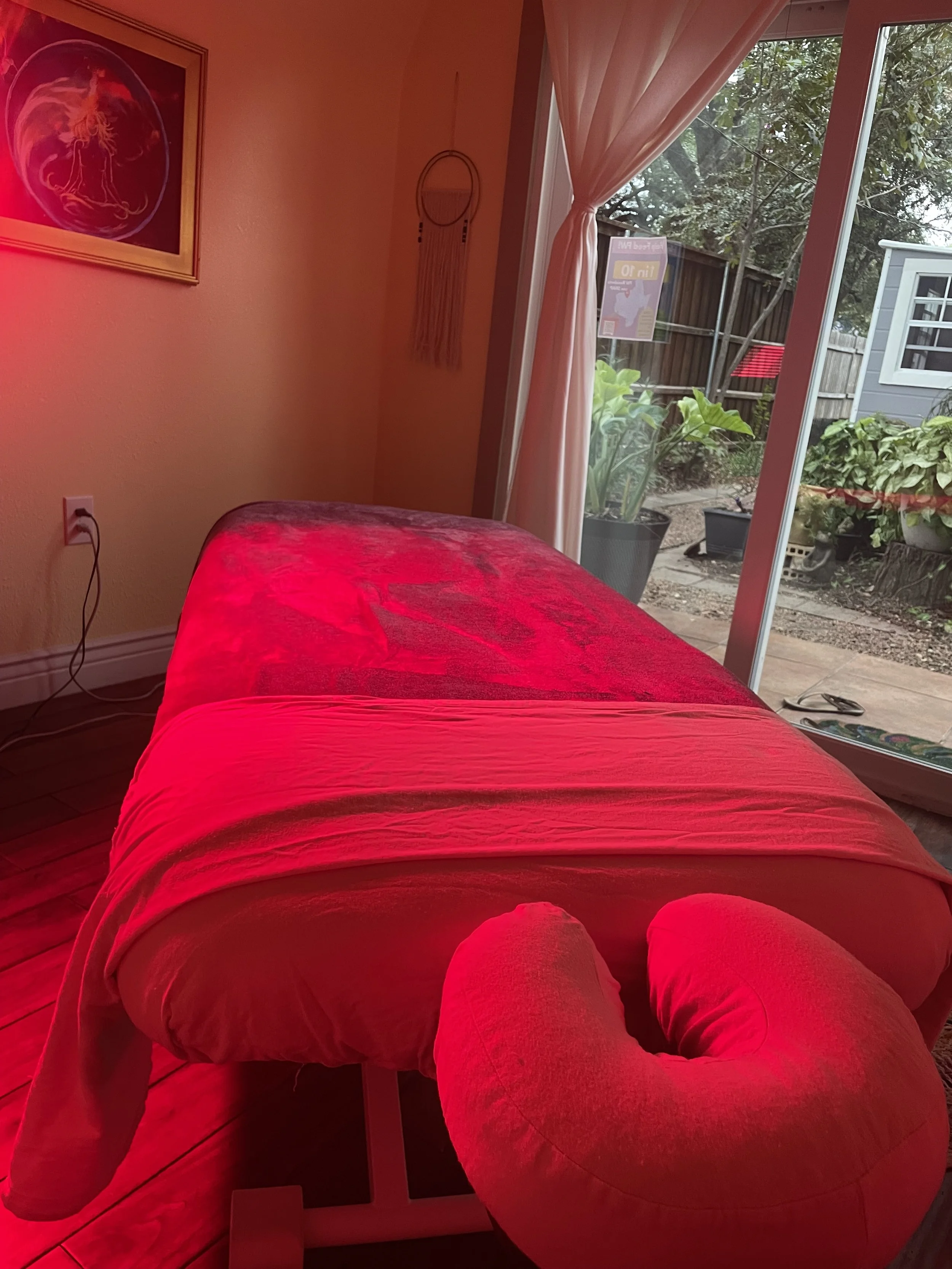 A massage or treatment table covered with a red blanket, with a red cushion at the head, inside a room with sliding glass door leading outside to a patio with plants.