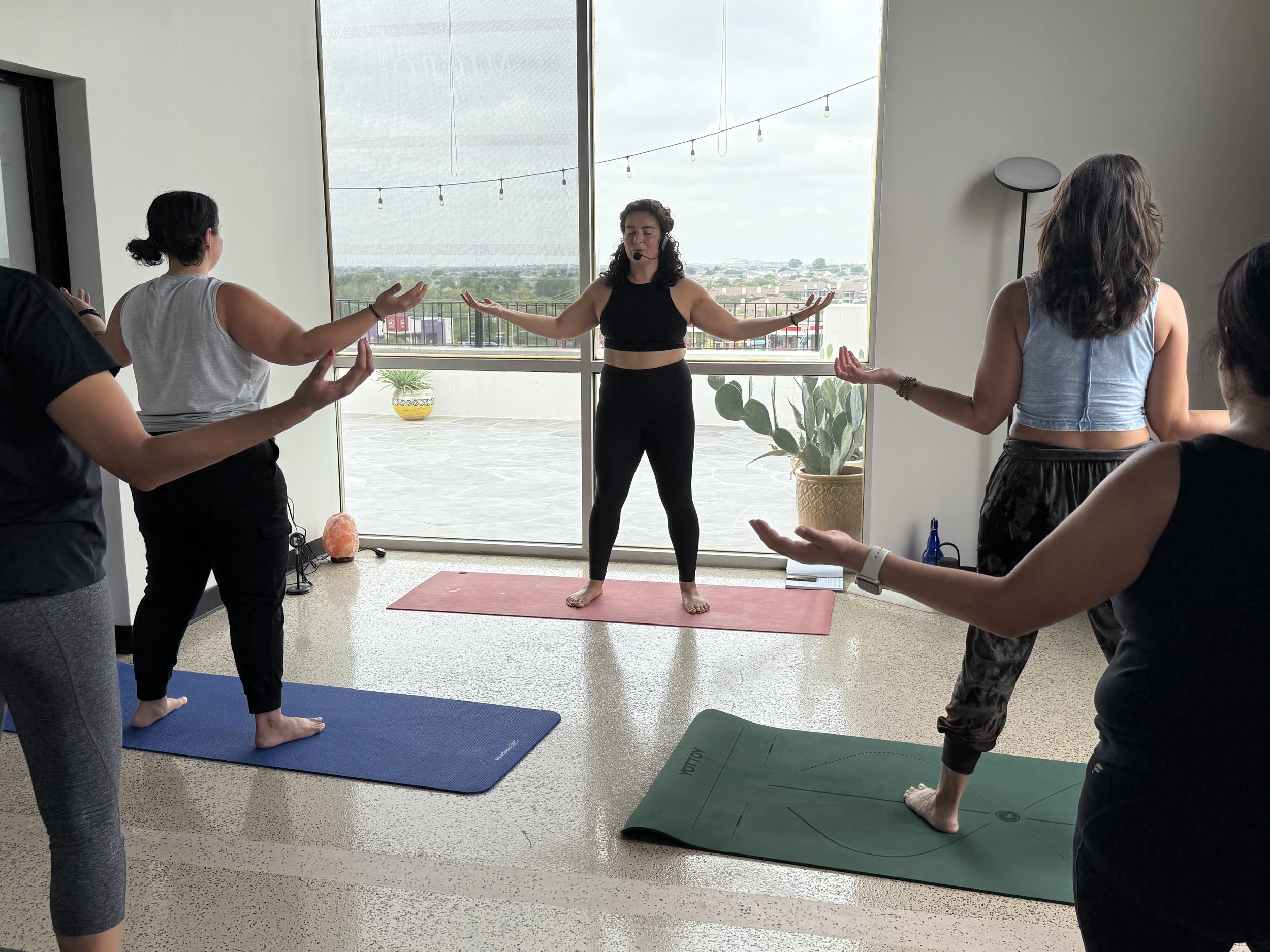A group of women participating in a yoga class indoors, with a teacher leading them in a breathing or meditation exercise near large windows overlooking an outdoor patio and cityscape.