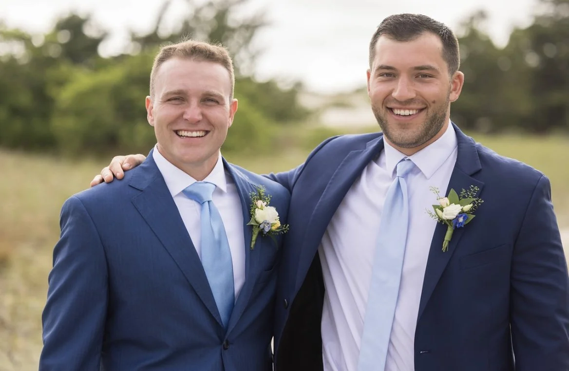 Two men in blue suits smiling outdoors, with one having his arm around the other's shoulder, both wearing boutonnières with flowers.