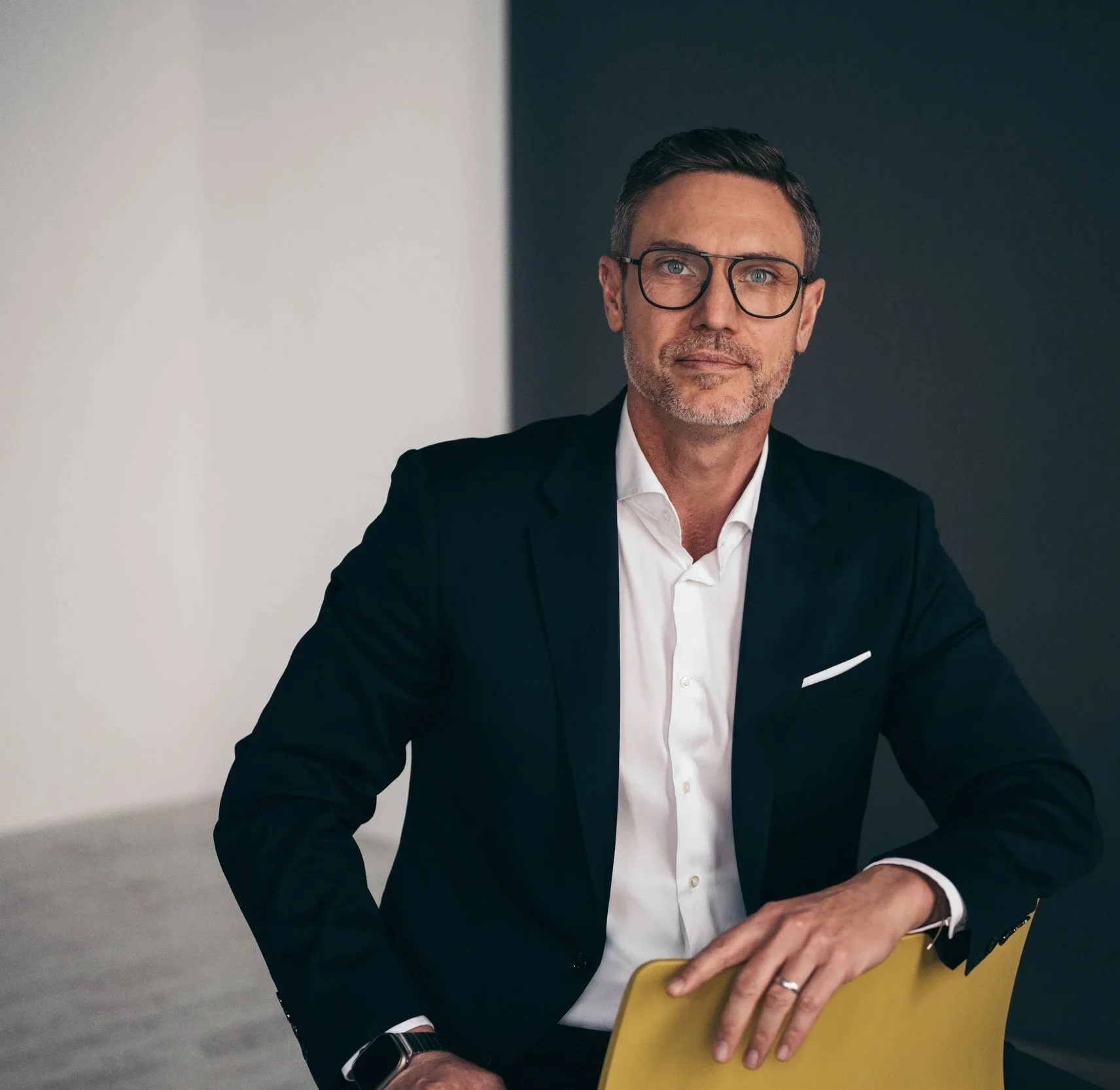 Ein Mann in einem schwarzen Anzug mit Brille sitzt an einem Tisch, hält eine gelbe Akte und schaut direkt in die Kamera.