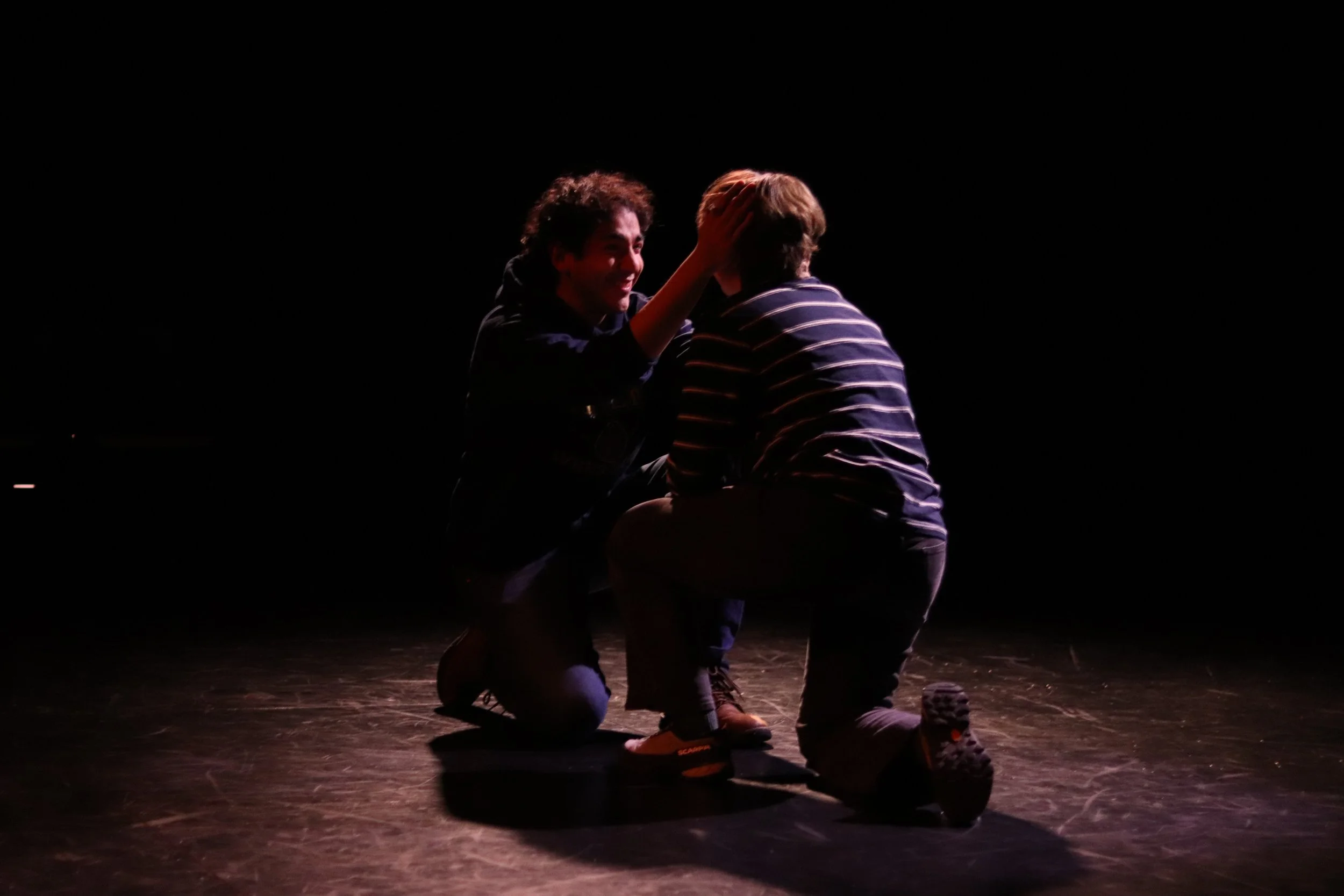 Two men on a dark stage, with one kneeling and holding the face of the other, who is also kneeling, with a spotlight focused on them.