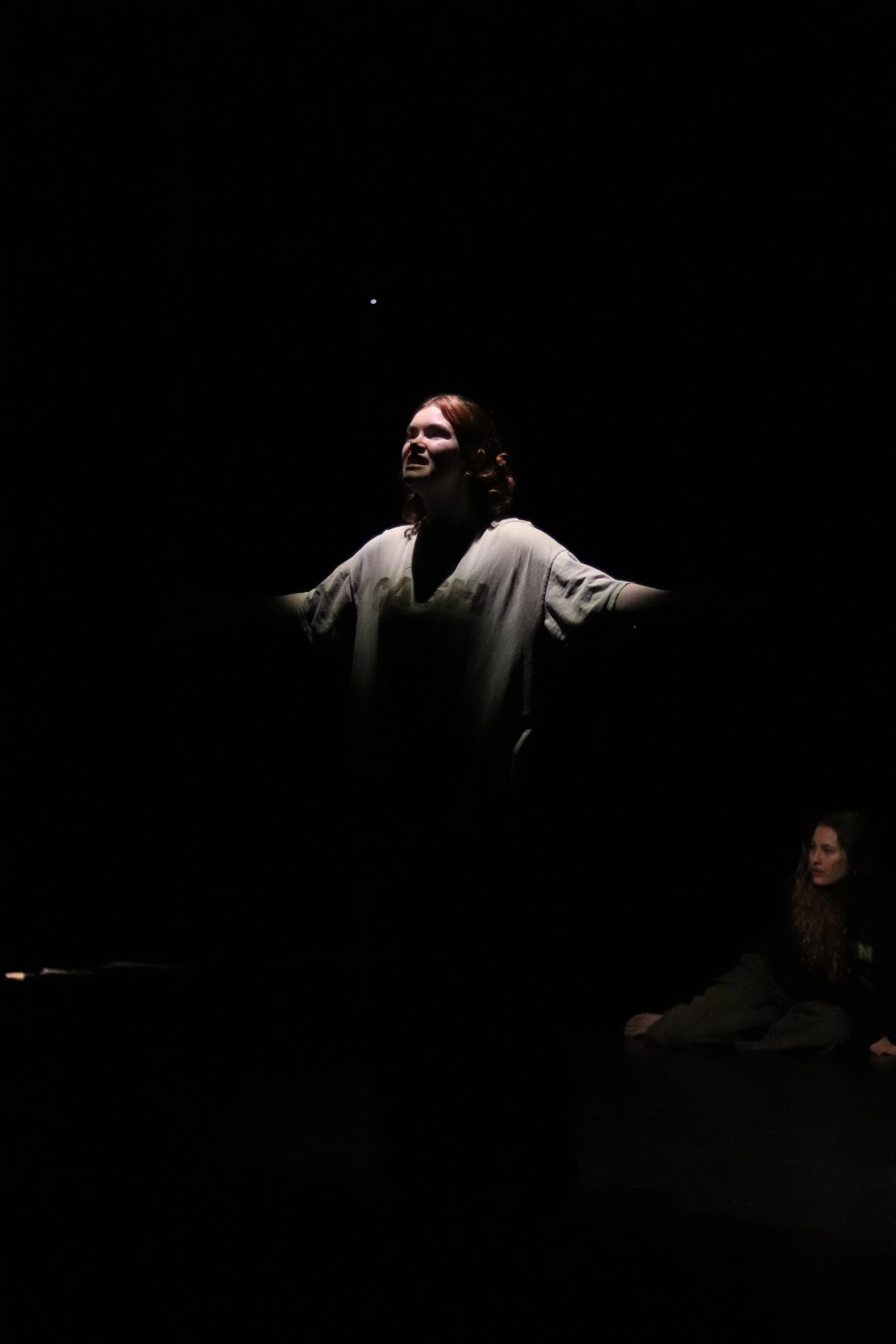 A woman with red hair standing with arms outstretched in a dark space, illuminated by a single light source above her, with another person sitting on the floor in the background.