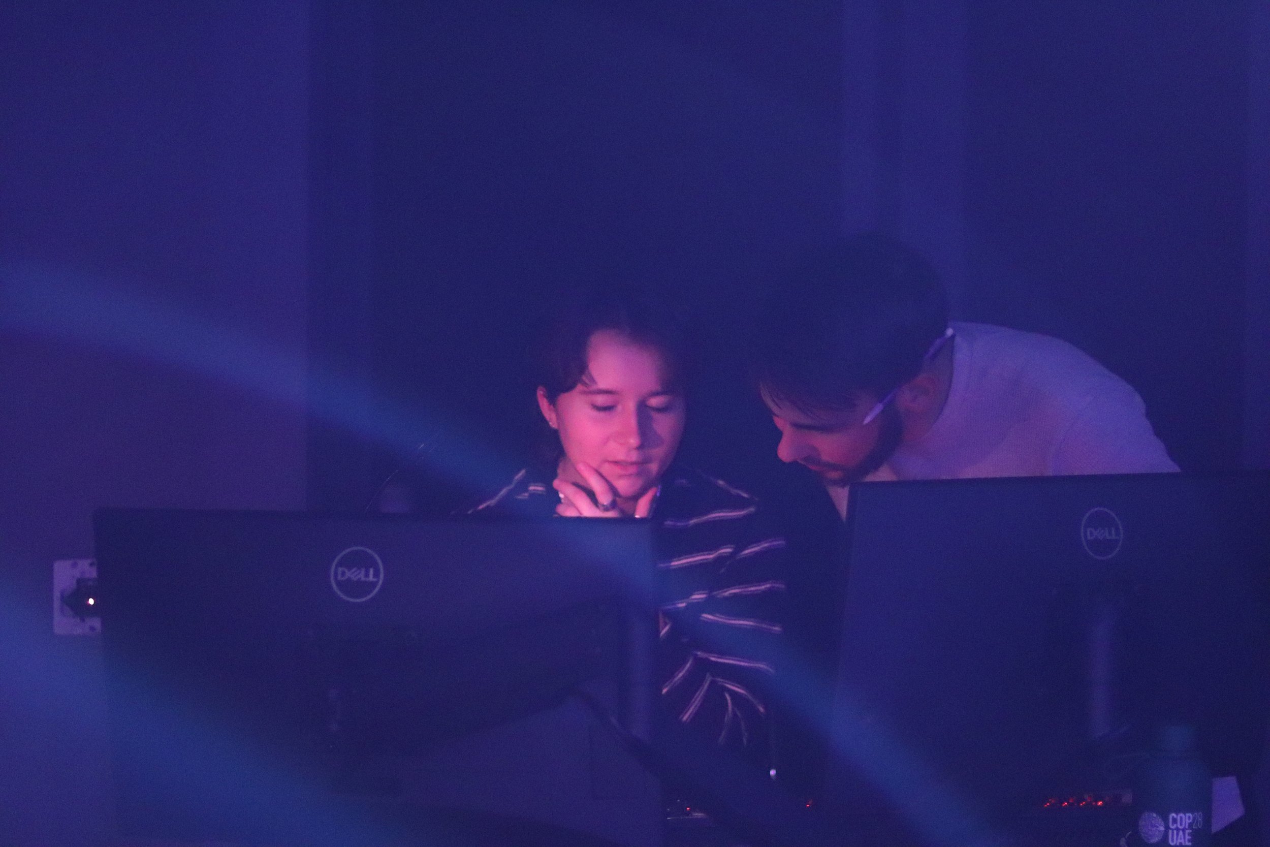 Two men in a dark room with purple lighting working on laptops.
