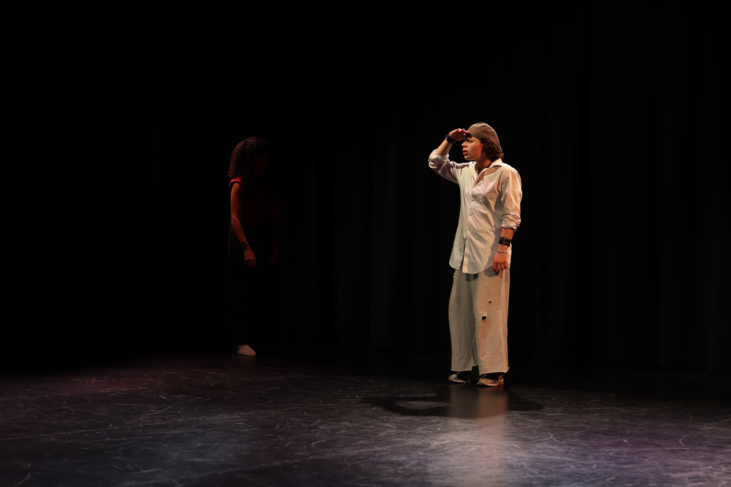 Two women on stage, one standing and shielding her eyes, the other in the background, with a black curtain backdrop.