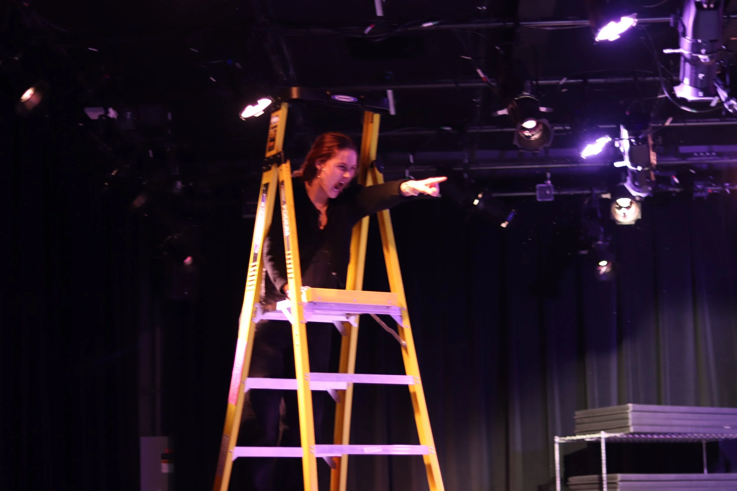 Woman standing on a ladder, pointing and shouting on stage with black curtains and lighting equipment.